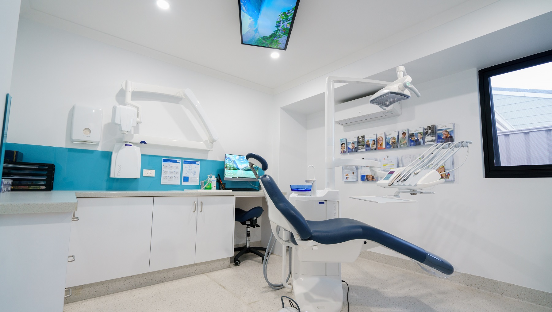 The walls of the examination room are white with sky-blue accents. The clinic has a reclining examination chair in the middle of the room where the basic equipment used by periodontists are attached, such as the bright light used during the examination, spittoon, and suction. Sunlight is visible through the glass window. There are magazine racks mounted on the wall on the right side of the room where reading materials are displayed. The walls of the examination room are white with sky-blue accents. The clinic has a reclining examination chair in the middle of the room where the basic equipment used by periodontists are attached, such as the bright light used during the examination, spittoon, and suction. Sunlight is visible through the glass window. There are magazine racks mounted on the wall on the right side of the room where reading materials are displayed.