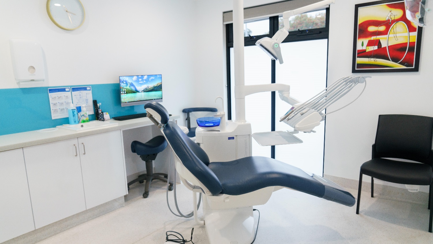 This view of an examination room shows a painting posted on the wall. The artist used bright and vibrant shades of yellow and red. It has a dark blue reclining examination chair that looks very comfortable. This chair is attached to the equipment used by dentists/periodontists. A wall clock is mounted on the wall and below it is a rectangular white tissue dispenser. A black chair is also kept near the examination chair and dental equipment. This view of an examination room shows a painting posted on the wall. The artist used bright and vibrant shades of yellow and red. It has a dark blue reclining examination chair that looks very comfortable. This chair is attached to the equipment used by dentists/periodontists. A wall clock is mounted on the wall and below it is a rectangular white tissue dispenser. A black chair is also kept near the examination chair and dental equipment.