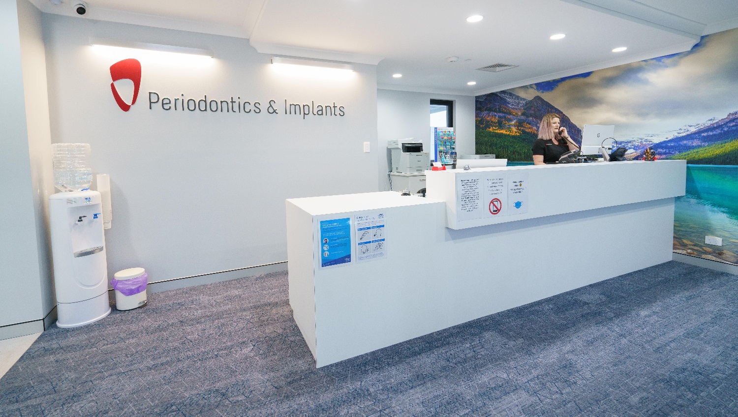 The long reception desk is painted white, the entire floor is carpeted, a large wall further to the right has a landscape picture of clouds, mountains, and water that adds a cozy feel to the room, and a white cabinet is placed at the back corner of the long desk while securing a space between them, to give more space for the clinic personnel. The clinic’s red logo is highlighted as its background wall is white. A desktop computer and telephone unit for use by the staff are on the desk. The long reception desk is painted white, the entire floor is carpeted, a large wall further to the right has a landscape picture of clouds, mountains, and water that adds a cozy feel to the room, and a white cabinet is placed at the back corner of the long desk while securing a space between them, to give more space for the clinic personnel. The clinic’s red logo is highlighted as its background wall is white. A desktop computer and telephone unit for use by the staff are on the desk.