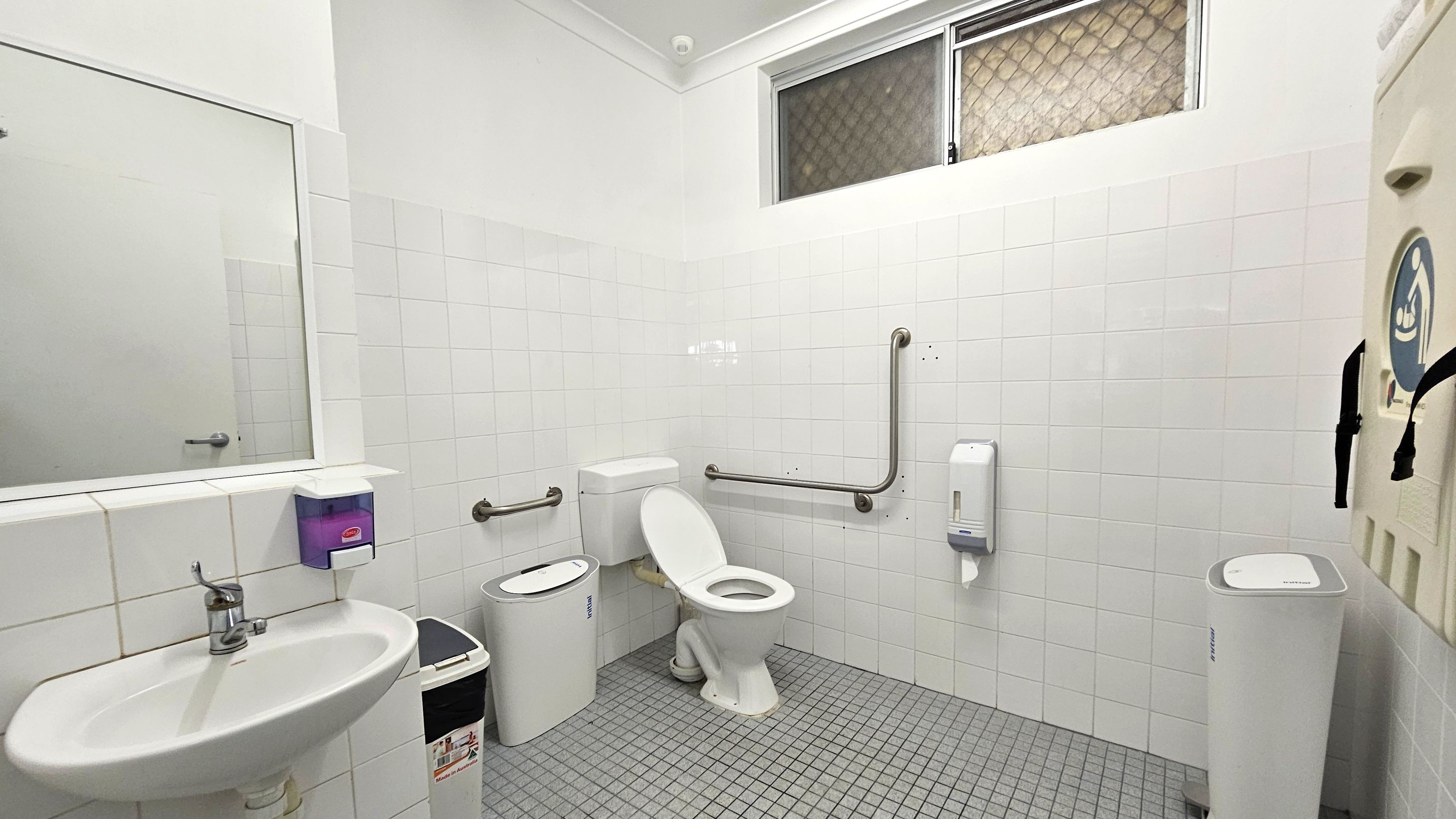 A public restroom with white tiled walls and a grey tiled floor. On the left, a sink with a mirror above it and a soap dispenser mounted on the wall. In the centre, a toilet with grab bars on both sides for accessibility. To the right, a nappy changing station is mounted on the wall, and a paper towel dispenser is nearby. A small window with a mesh screen is located high on the wall.