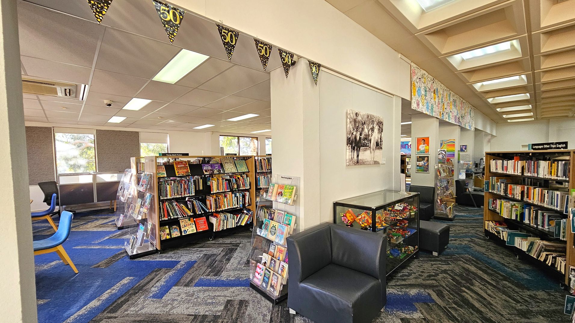 A library interior with a blue and grey patterned carpet. On the left, there are bookshelves filled with books and a few blue chairs near windows. In the centre, a display rack holds various books and magazines. A black armchair is positioned next to a glass display case containing colourful items. On the right, more bookshelves are labelled "Languages Other Than English." The ceiling features recessed lighting and decorative banners with "50" hang across the room.