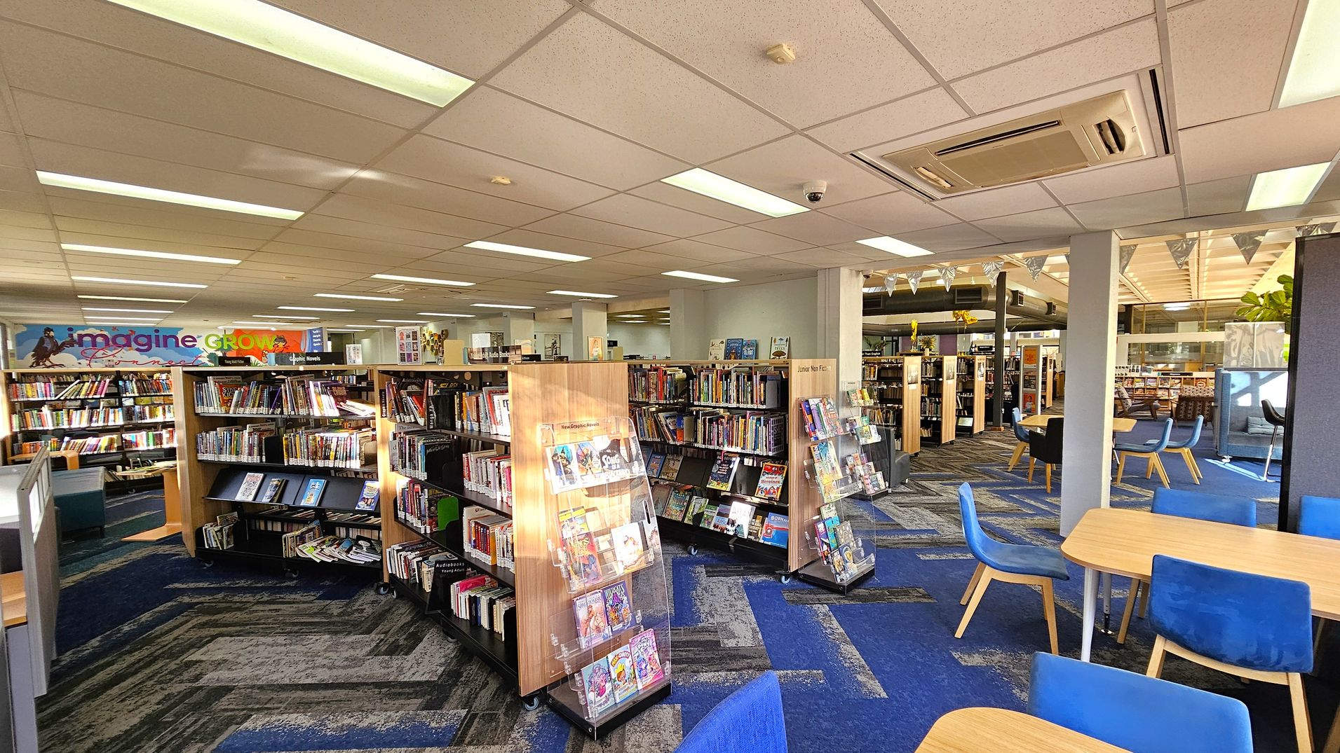 A library interior with bookshelves filled with books in the centre and left. A colourful sign with the words "imagine" and "GROW" is visible on the left wall. On the right, there are tables with blue chairs. The floor is carpeted in a blue and grey pattern, and the ceiling has recessed lighting and an air conditioning unit. In the background, more bookshelves and seating areas are visible, along with some decorative bunting.