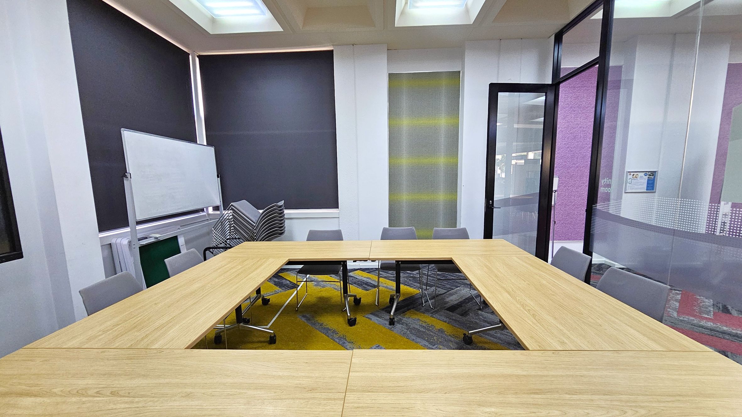 A meeting room with a U-shaped wooden table surrounded by grey chairs. On the left, a whiteboard is positioned against the wall, with a stack of chairs nearby. The walls have large windows covered with dark blinds. On the right, a glass door is open, leading to another area. The floor is carpeted with a geometric pattern in grey and yellow. The ceiling features recessed lighting panels.