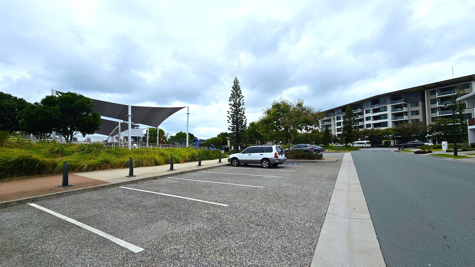 A parking area with a few cars is on the right, adjacent to a road. On the left, there is a grassy area with trees and a structure with large, triangular shade sails. In the background, a multi-storey building with balconies is visible. A tall, narrow tree stands in the centre, surrounded by other greenery. The ground is paved with asphalt, and the sky is overcast.