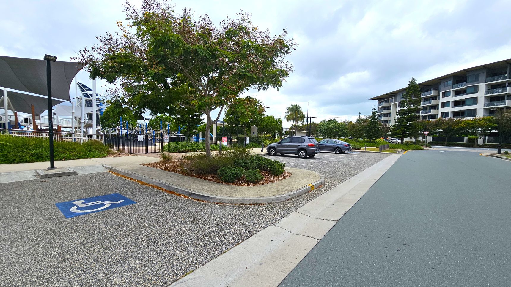 A parking area with a designated disabled parking space marked in blue on the left. A tree with green foliage is in the centre, surrounded by a small landscaped area. Two cars are parked on the right. In the background, there is a building with multiple floors and balconies. A playground with shade structures is visible on the left. The ground is paved with a mix of grey and green surfaces.