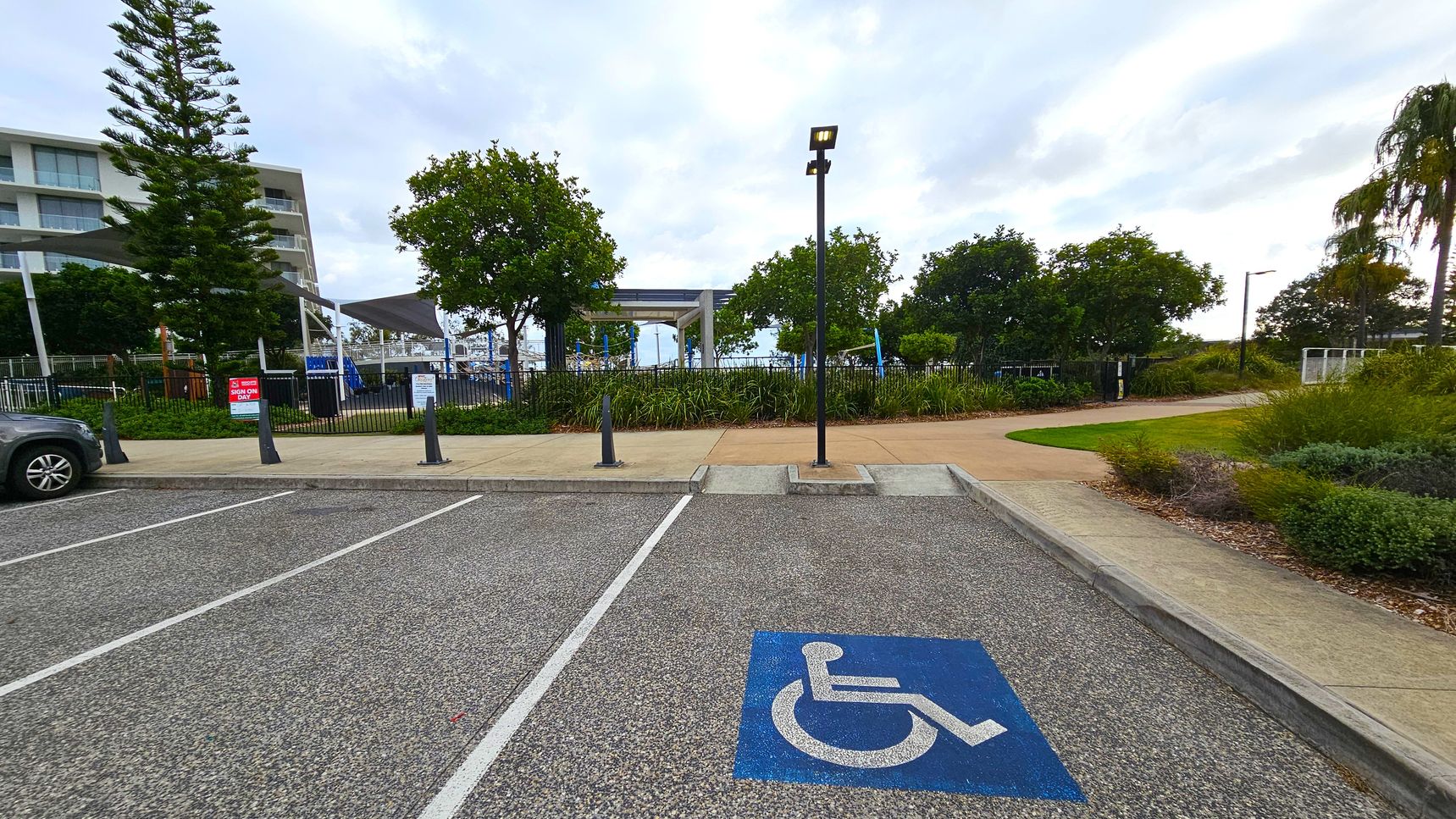 A car park with a designated disabled parking space marked by a blue symbol on the ground. On the left, a partial view of a car is visible. In the background, there are trees and a building with multiple floors. A pathway runs parallel to the parking area, bordered by grass and shrubs. A tall lamp post stands near the centre, and a sign is visible near the entrance to a fenced area. The ground is paved with a textured surface.