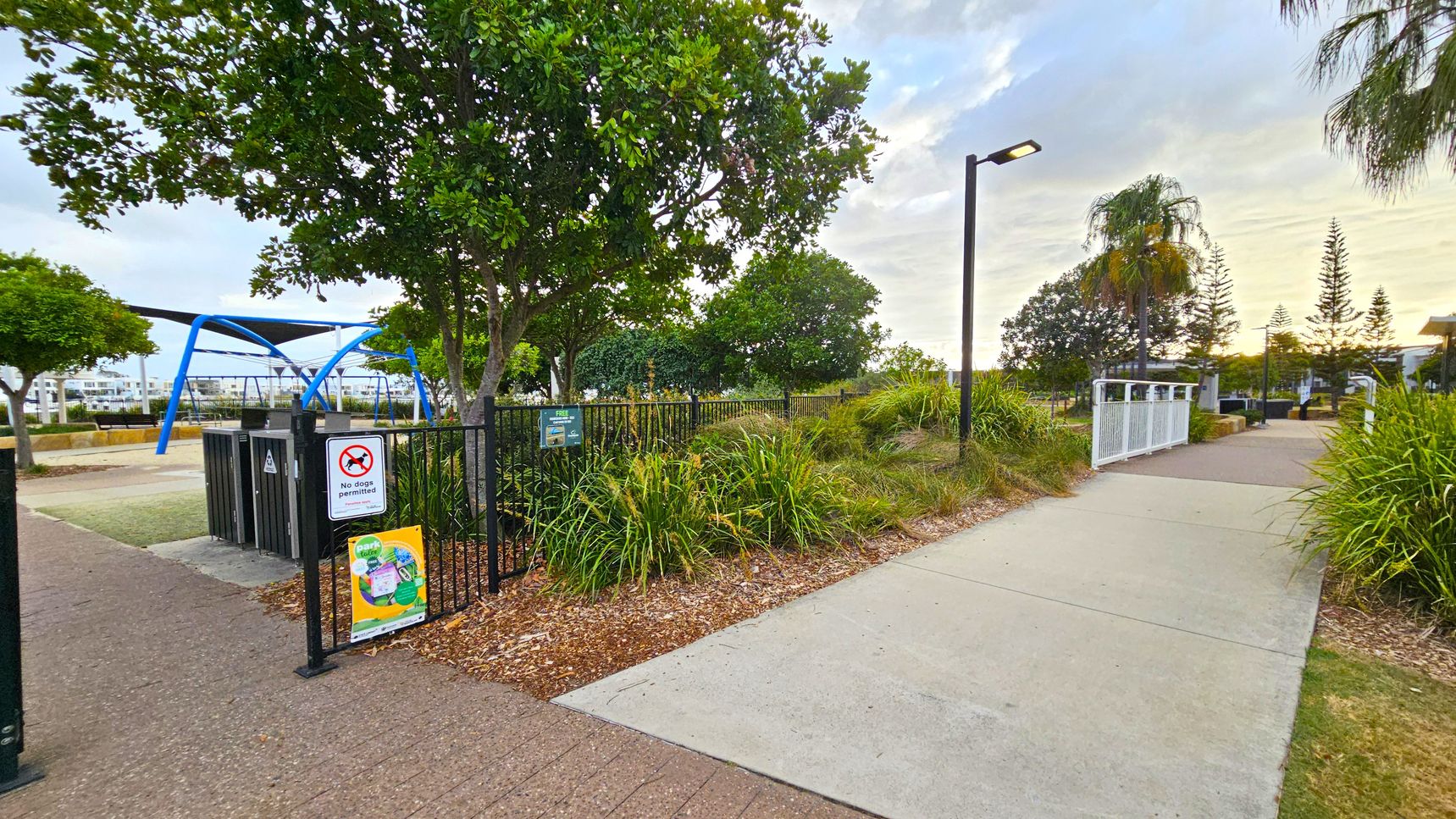 A park scene with a paved path on the right leading to a white gate. On the left, there's a fenced playground area with a "No dogs permitted" sign and a colourful poster. Blue play equipment is visible behind the fence. Trees and shrubs are scattered throughout, with a lamp post along the path. In the background, there are more trees and some buildings. The ground is covered with mulch and grass.