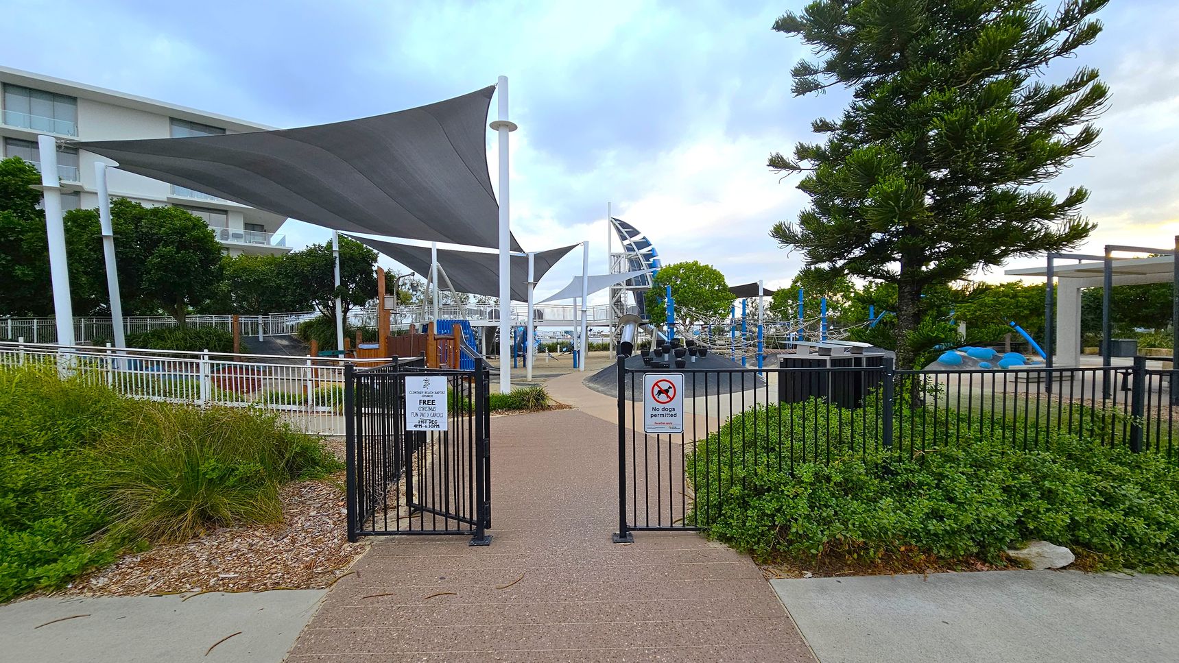 A pathway leads through an open gate into a playground area. On the left, there are white railings and lush greenery. Large triangular shade sails are suspended above, supported by tall white poles. In the centre, playground equipment is visible, including climbing structures. On the right, a tall tree stands next to more playground apparatus. Signs on the gate indicate rules, including "No dogs permitted." A modern building is partially visible in the background.