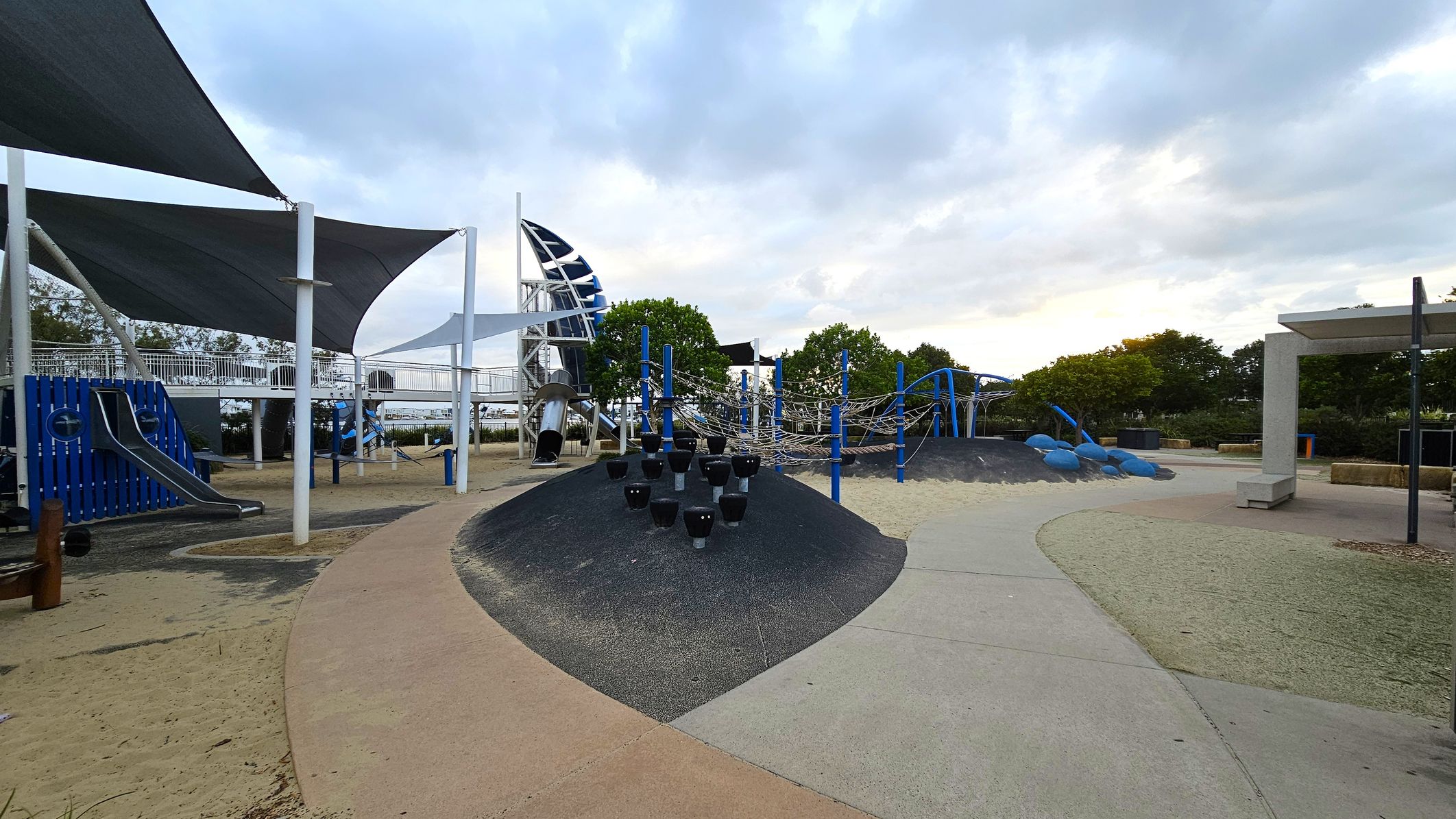 A playground with various equipment. On the left, there are blue and silver slides under large triangular shade sails supported by white poles. In the centre, a black mound with small climbing structures and a rope climbing frame with blue supports. On the right, a path leads to a shaded seating area. The ground is a mix of sand and rubber surfaces, and there are trees in the background.