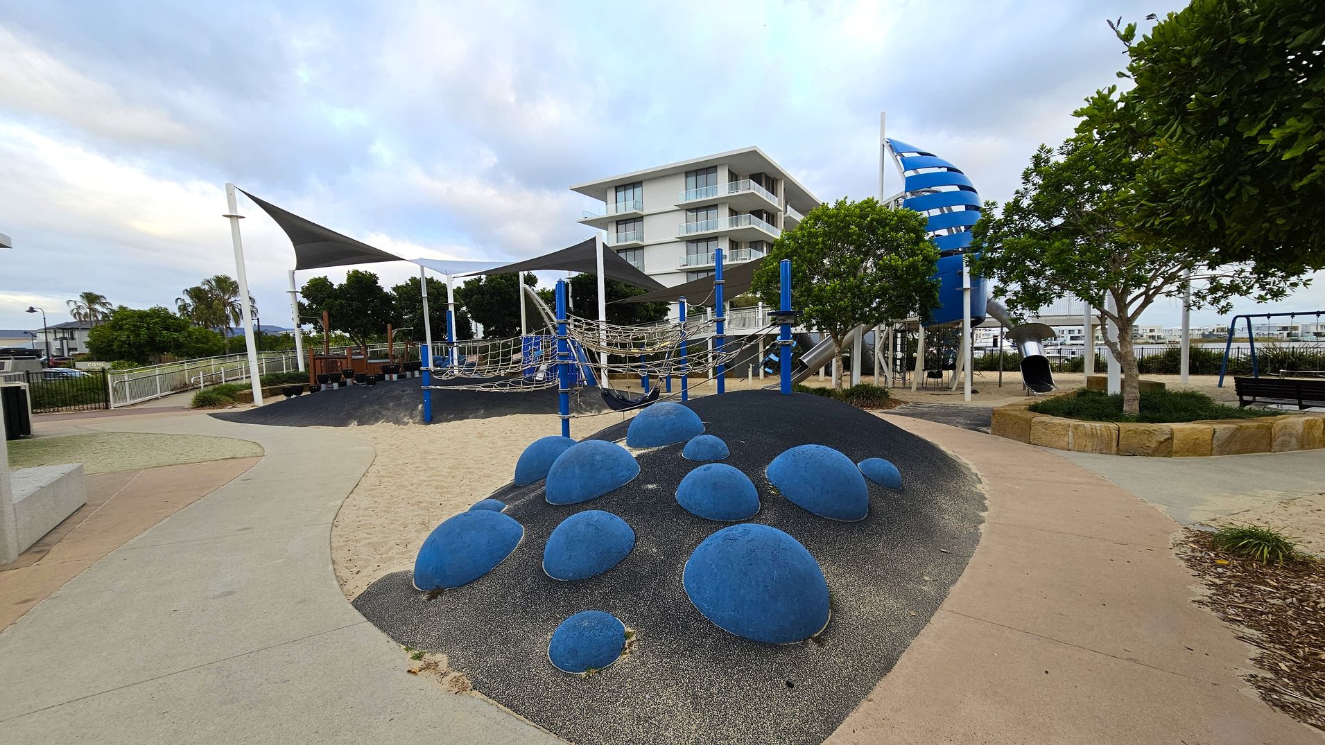 A playground with a sandy surface and a path winding through it. On the left, there are shade sails over a climbing structure with ropes and blue poles. In the centre, a mound with blue dome-shaped climbing features. On the right, a tall blue spiral slide and swings. Trees are scattered throughout, and a modern white building is visible in the background. The ground is a mix of sand, rubber, and concrete.