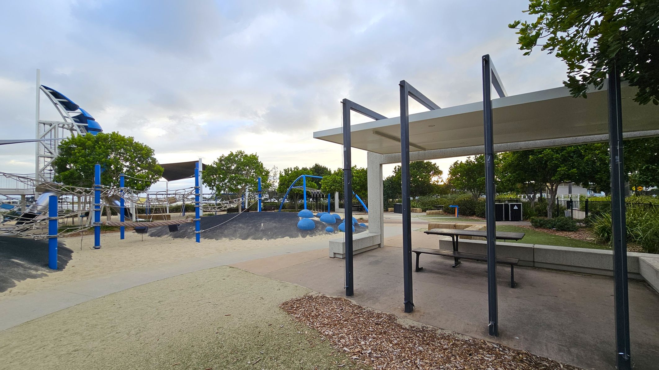 A playground with a variety of equipment. On the left, there is a climbing structure with blue and white elements, including nets and slides. In the centre, blue climbing rocks are visible on a mound. On the right, a sheltered area with a picnic table and bench is present. The ground is a mix of sand and rubber surfaces. Trees and shrubs are scattered throughout, with a fence and more greenery in the background.