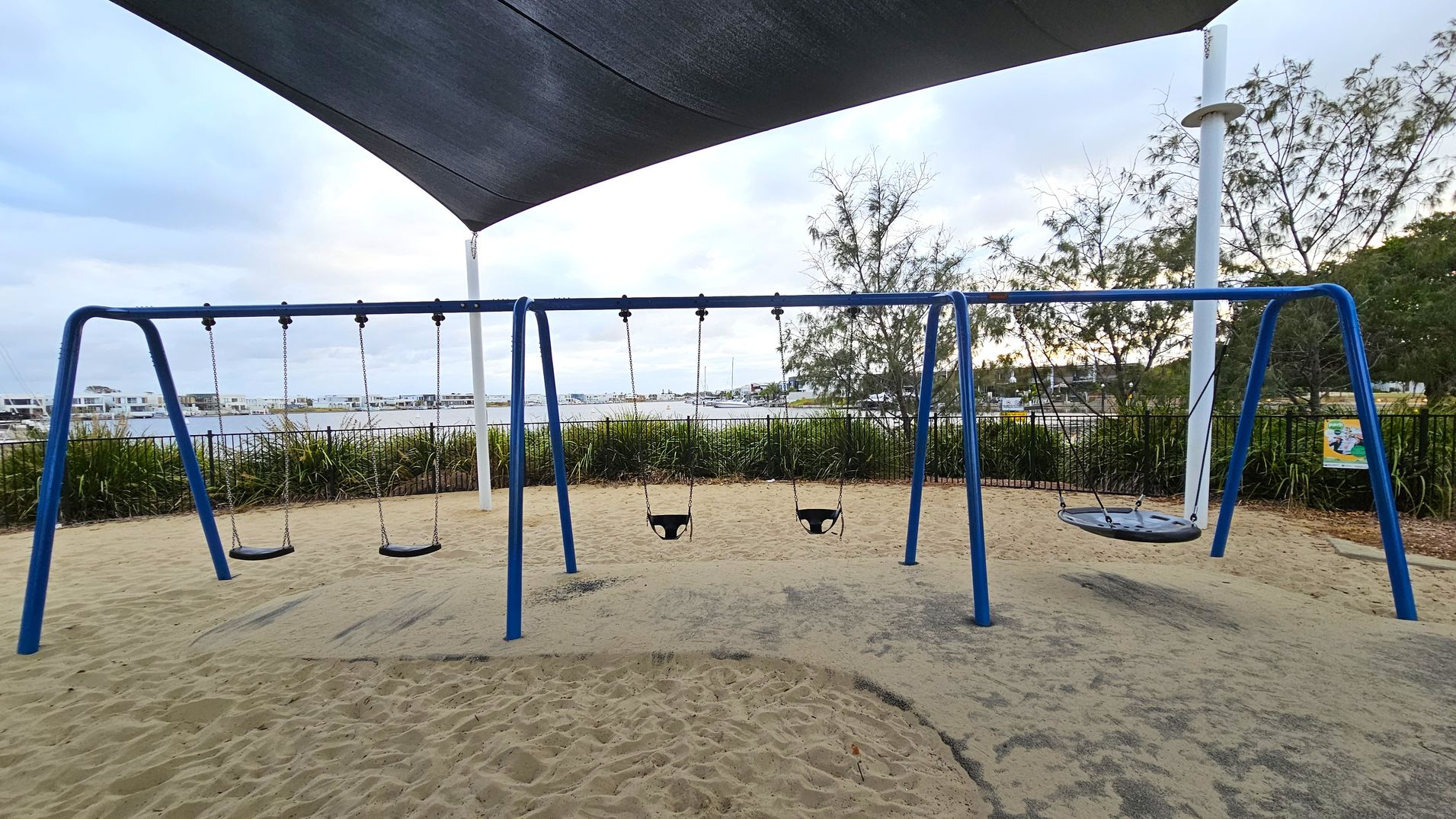 A playground with a blue swing set featuring four swings: two regular swings, two baby swings, and a circular net swing. The ground is covered in sand with patches of green matting. In the background, there is a body of water with boats and a line of modern buildings. Trees and shrubs are visible along a fence, and a large shade sail is overhead.