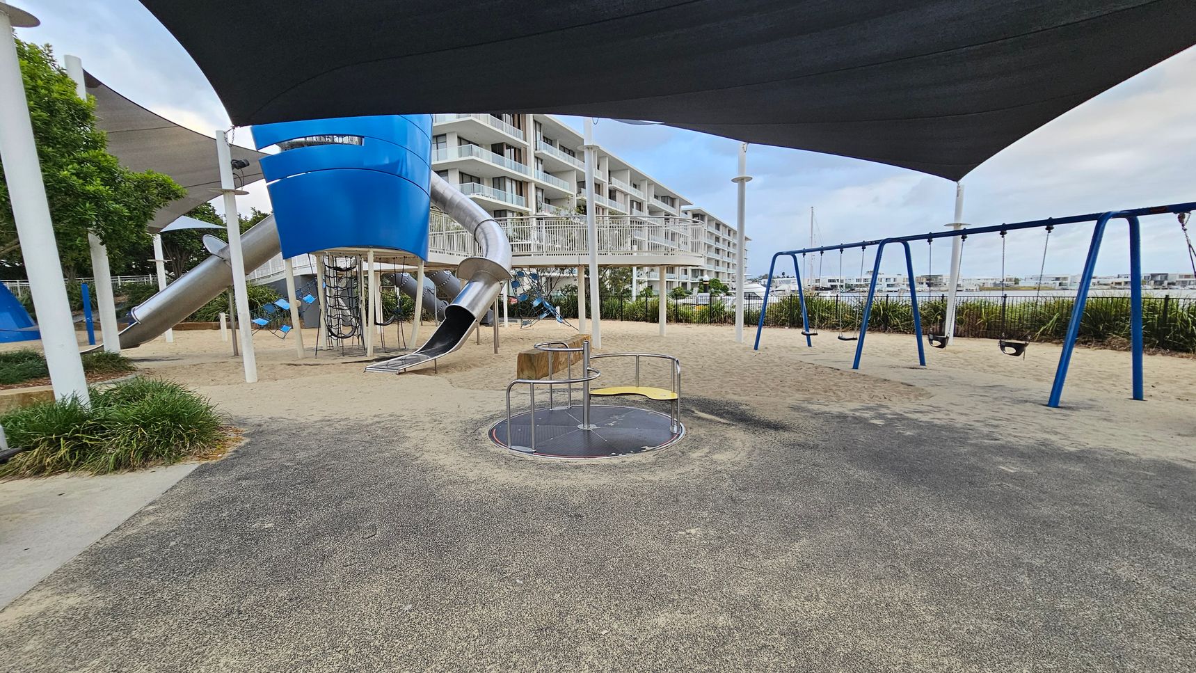 A playground with a blue and silver slide structure on the left, featuring a climbing net and a tube slide. In the centre, there's a roundabout with metal bars and a yellow seat. On the right, a blue swing set with several swings. The ground is covered with a mix of sand and green rubber matting. Overhead, there are large black shade sails. In the background, a multi-storey building with balconies is visible, along with some greenery and a fence.