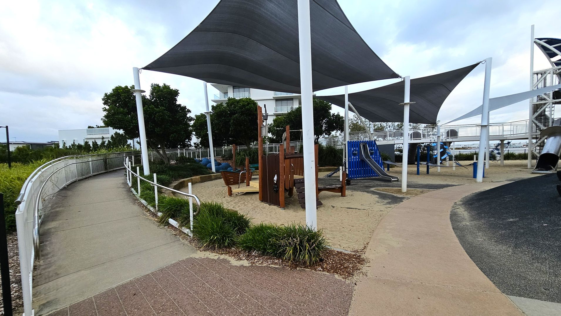 A playground with a sandy area in the centre featuring wooden play structures. To the right, there is a blue slide and a large metal slide. Overhead, several large shade sails are supported by white poles. On the left, a curved path with a white railing runs alongside greenery. In the background, there are trees and a white building. The ground is a mix of sand, concrete, and rubberised surfaces.