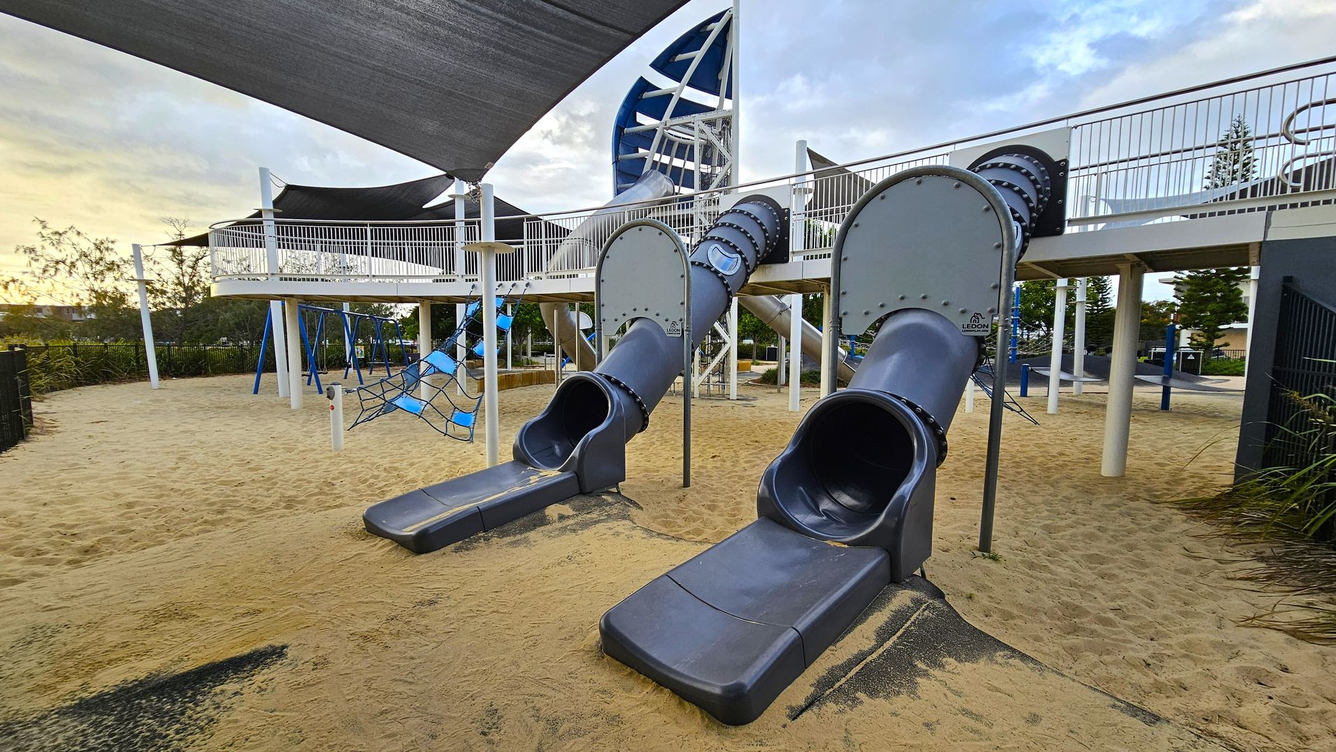 A playground with two large tube slides in the centre, emerging from an elevated platform supported by white pillars. The platform is covered with a black shade sail. To the left, there's a climbing net structure and swings. The ground is covered with sand, and there are trees and bushes in the background. The playground equipment is primarily blue and grey.