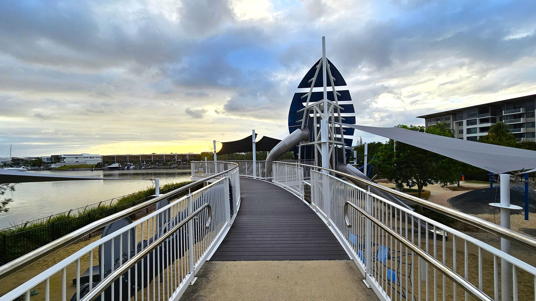 A walkway with white railings leads towards a modern, abstract structure resembling a sail. The path is made of dark planks, contrasting with the light-coloured ground. On the left, a body of water with moored boats is visible, bordered by grass. On the right, there are trees and a multi-storey building. The sky is overcast with patches of blue and grey clouds.