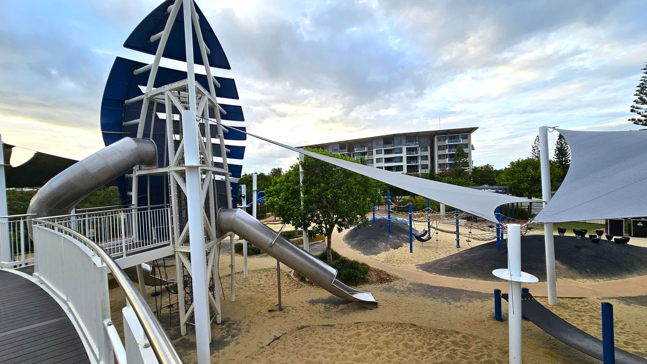 A playground with a large climbing structure on the left, featuring a metal slide and blue accents. The ground is covered in sand. In the middle, there are white shade sails and a tree. On the right, a building with multiple floors is visible in the background, along with more playground equipment, including ropes and a hammock. The area is surrounded by greenery and a few tall trees.
