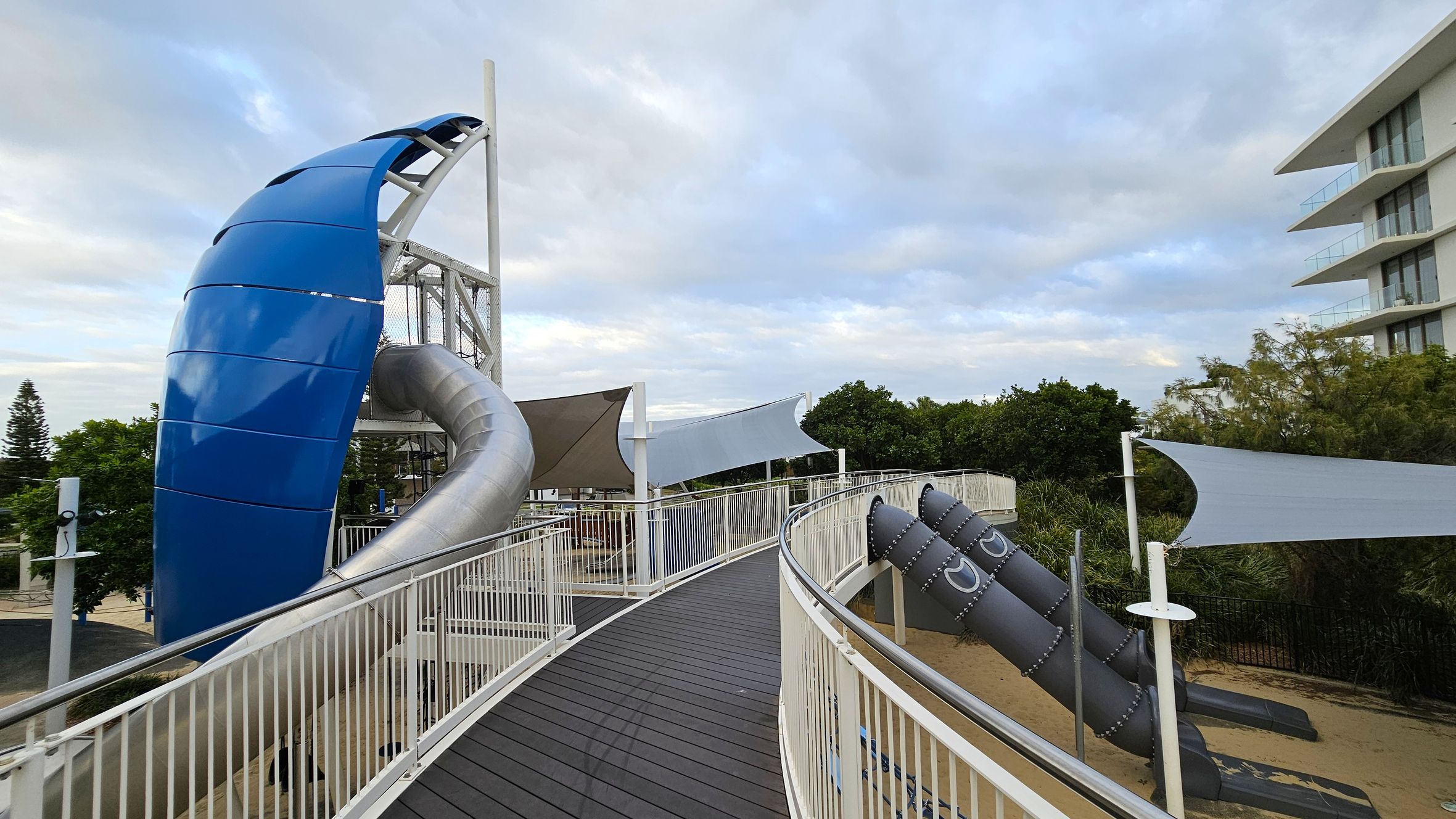 A playground with a large blue and silver slide structure on the left, featuring a curved design. A walkway with white railings leads towards the centre, with shade sails overhead. On the right, there are two dark grey tube slides with rivet details, set over a sandy area. Trees and a multi-storey building are visible in the background.