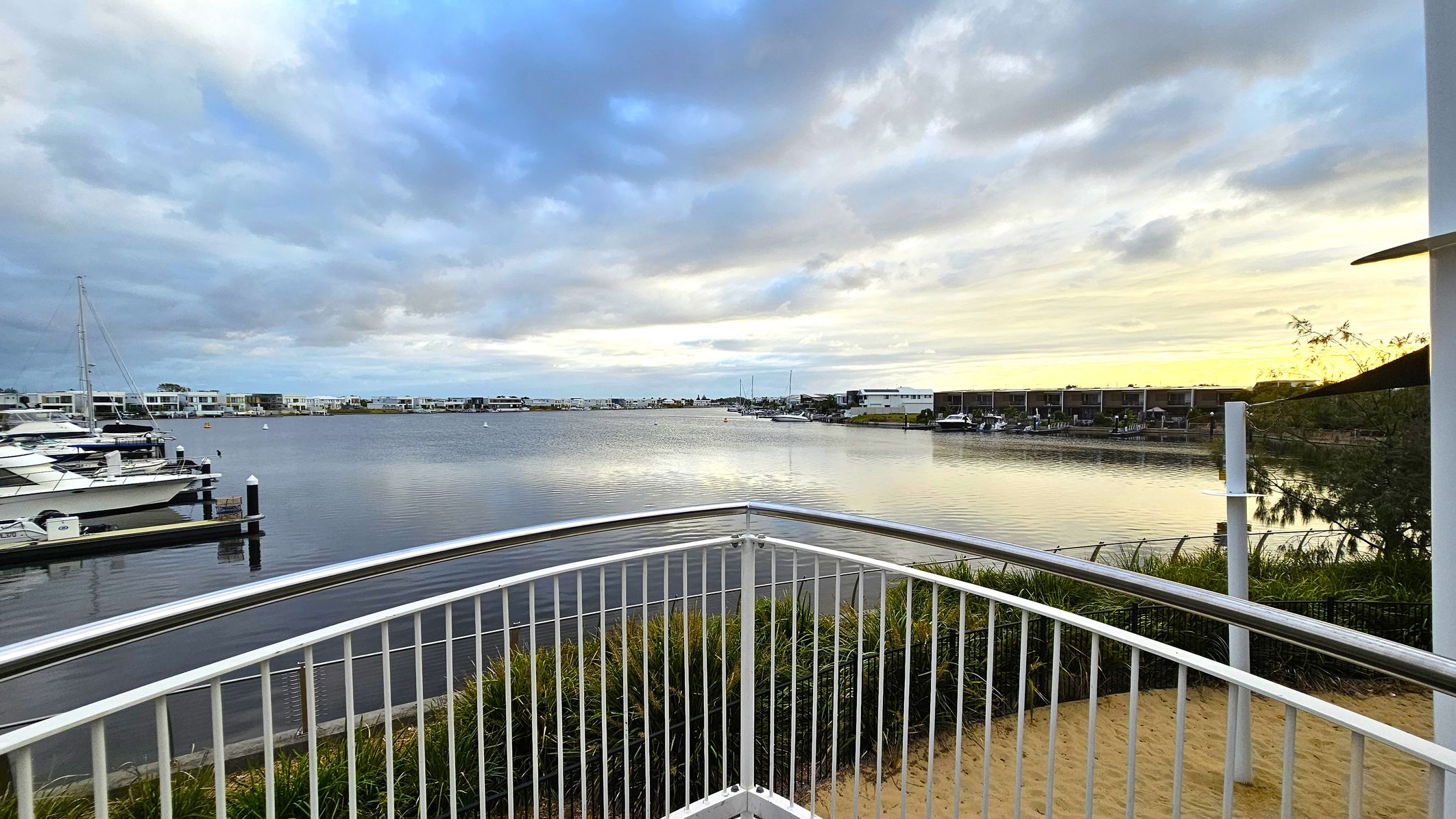 A waterfront scene with a metal railing in the foreground, overlooking a calm body of water. On the left, several boats are docked at a marina. In the background, modern houses line the water's edge, with more boats visible. The right side features some greenery and a white post. The sky is overcast with patches of light breaking through, reflecting on the water's surface.