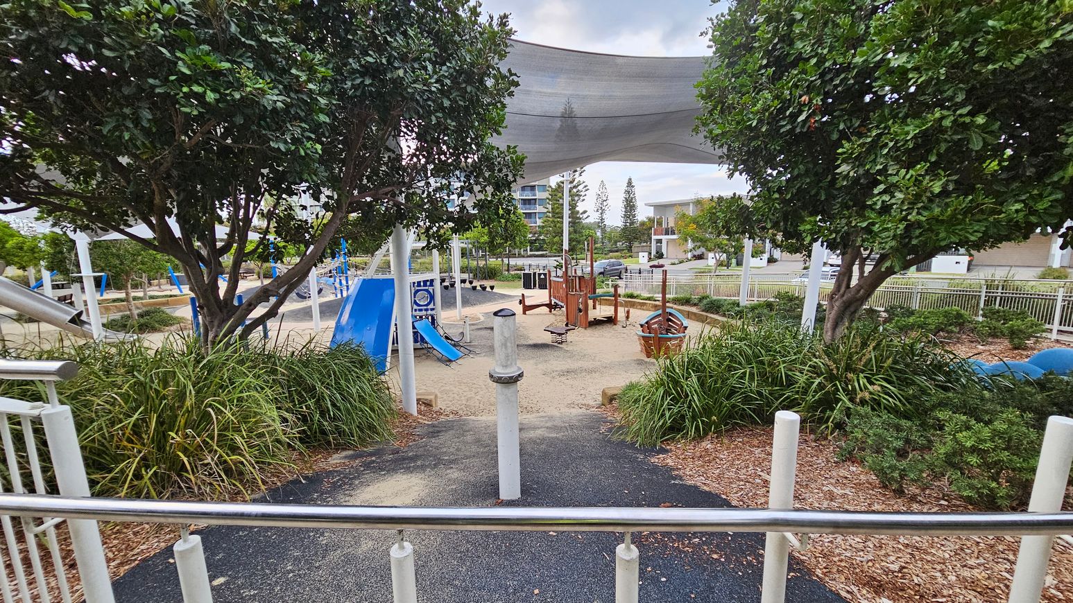 A playground with a sandy area in the centre, featuring a wooden play structure and a blue slide. Two large trees with dense foliage are on the left and right, surrounded by green shrubs. A shade sail is overhead, providing cover. The foreground has a path with a metal railing and a post. In the background, there are buildings and more trees, with a fence enclosing the playground.