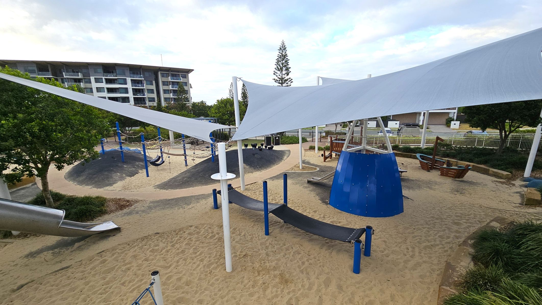 A playground with sandy ground and various play structures. In the foreground, there are blue and black climbing and balancing equipment. White shade sails are stretched overhead. In the background, a large blue cone-shaped structure and a slide are visible. Trees and shrubs are scattered around, and a multi-storey building is in the distance. A pathway winds through the area, and there are benches and additional play equipment.