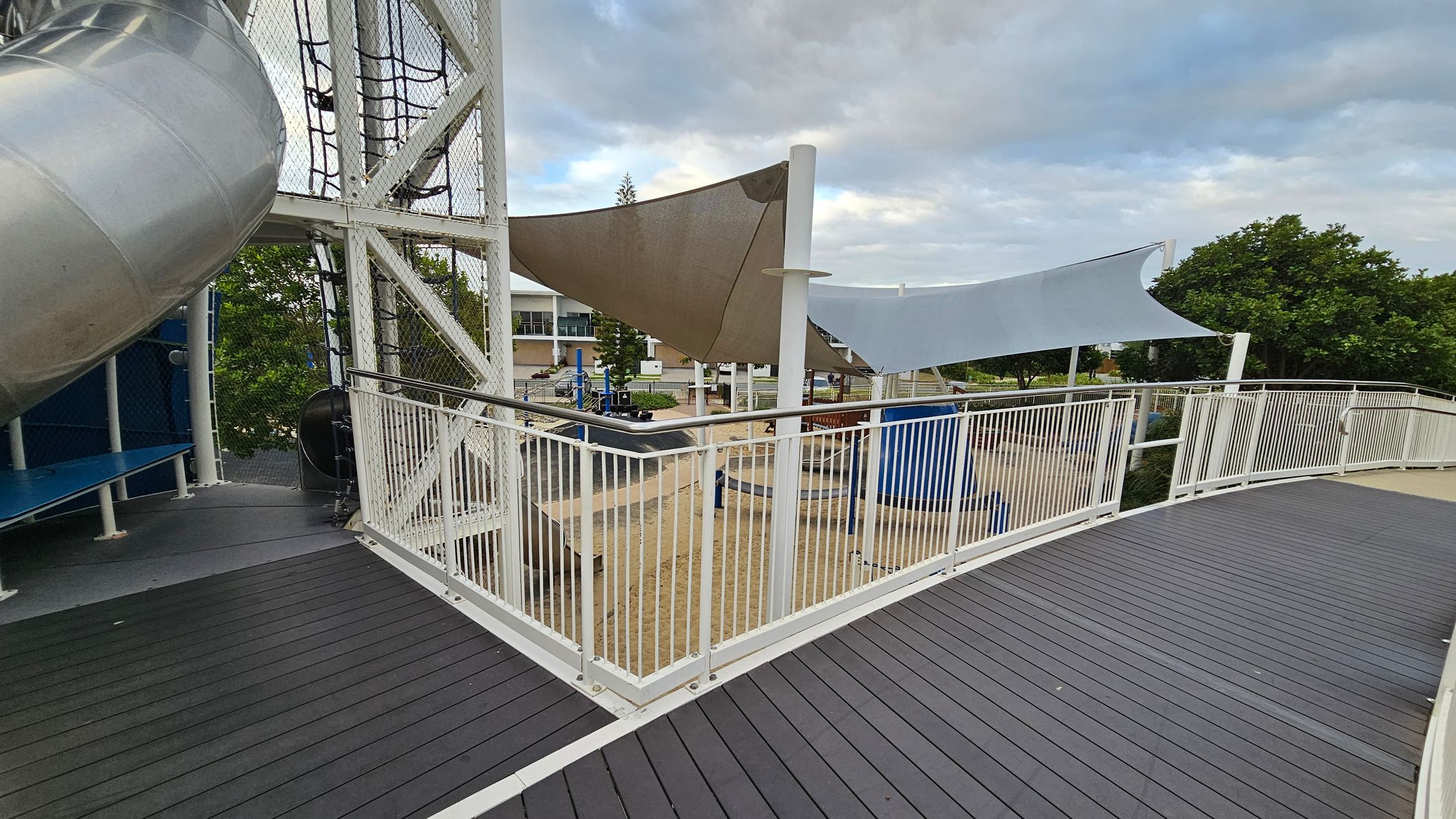 A playground area with a large metal slide on the left, supported by a white metal structure with netting. The ground is covered with dark grey decking. In the centre, there are triangular shade sails in beige and light blue, supported by white poles. Below, a sandy play area with various equipment is visible. Trees and a building are in the background, with a white railing running along the walkway.