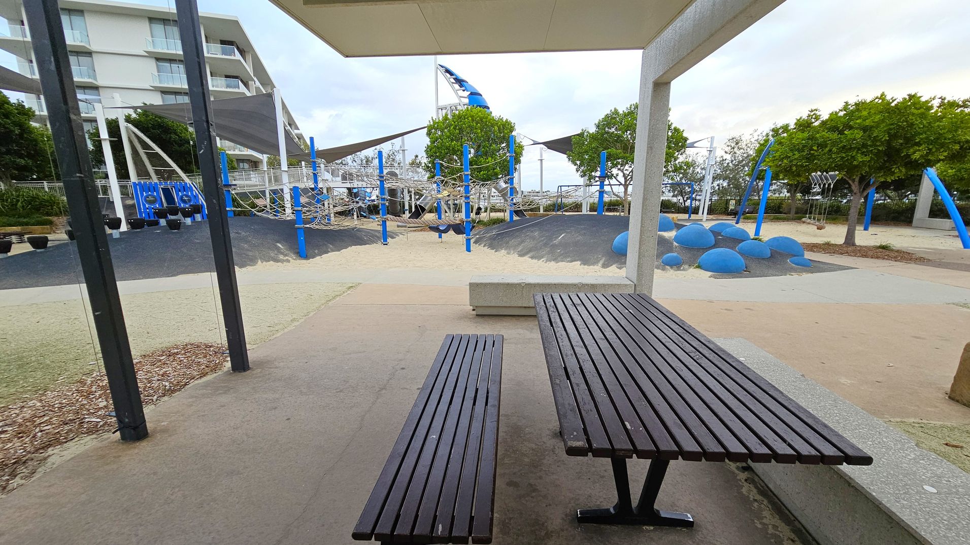 A playground with a focus on a picnic area in the foreground, featuring a black metal bench and table on a concrete surface. In the middle, there are climbing structures with blue poles and ropes, and a mound with blue dome-shaped features. The background includes trees and a modern building with balconies. The ground is a mix of sand, mulch, and rubberised surfaces. A shade structure is partially visible above the playground.