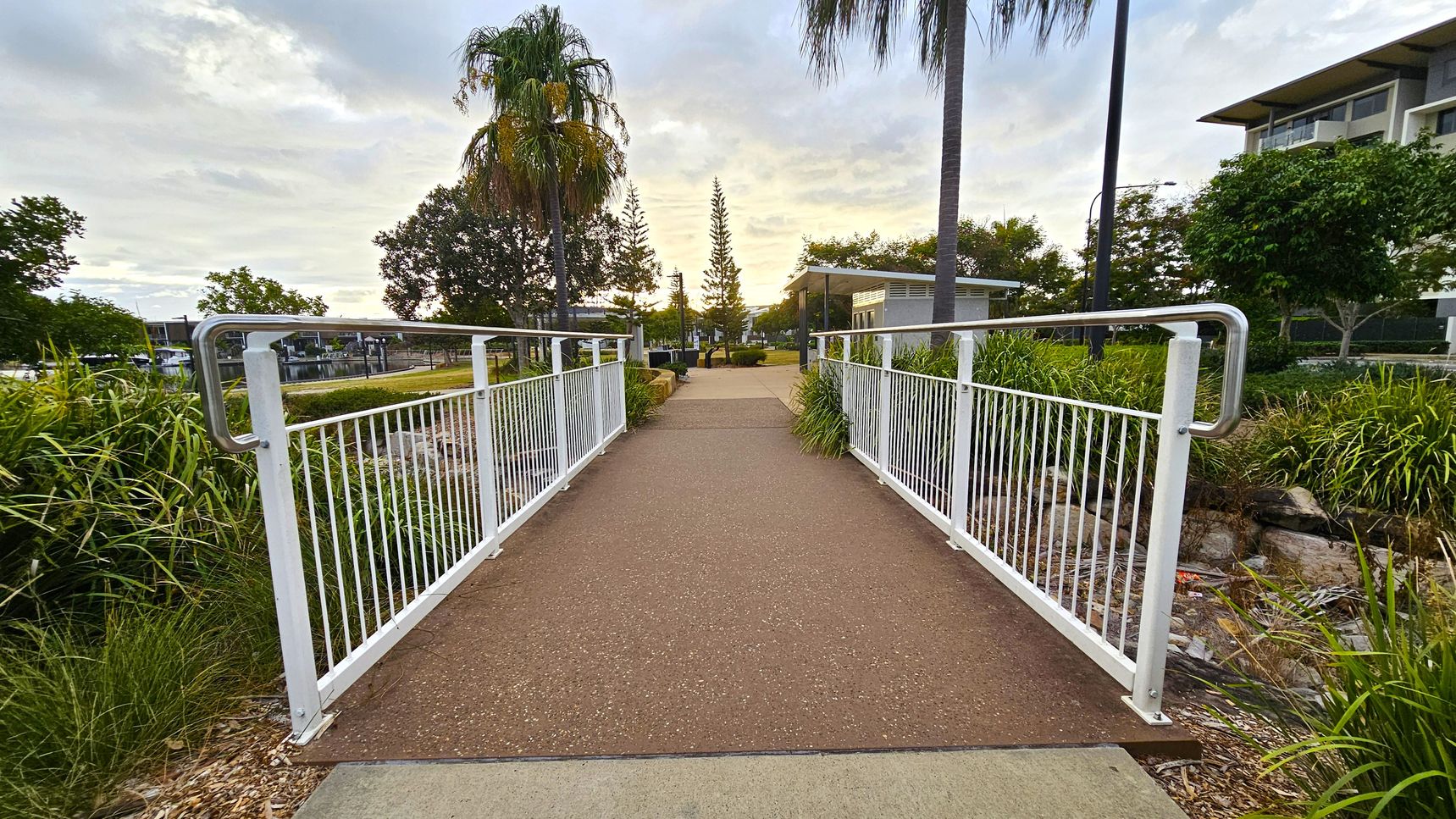 A pathway with a reddish-brown surface leads over a small bridge with white railings. On either side, there are green plants and grass. In the background, there are tall trees, including a palm tree on the left and a conifer in the centre. To the right, a modern building is partially visible. The sky is overcast with light clouds.