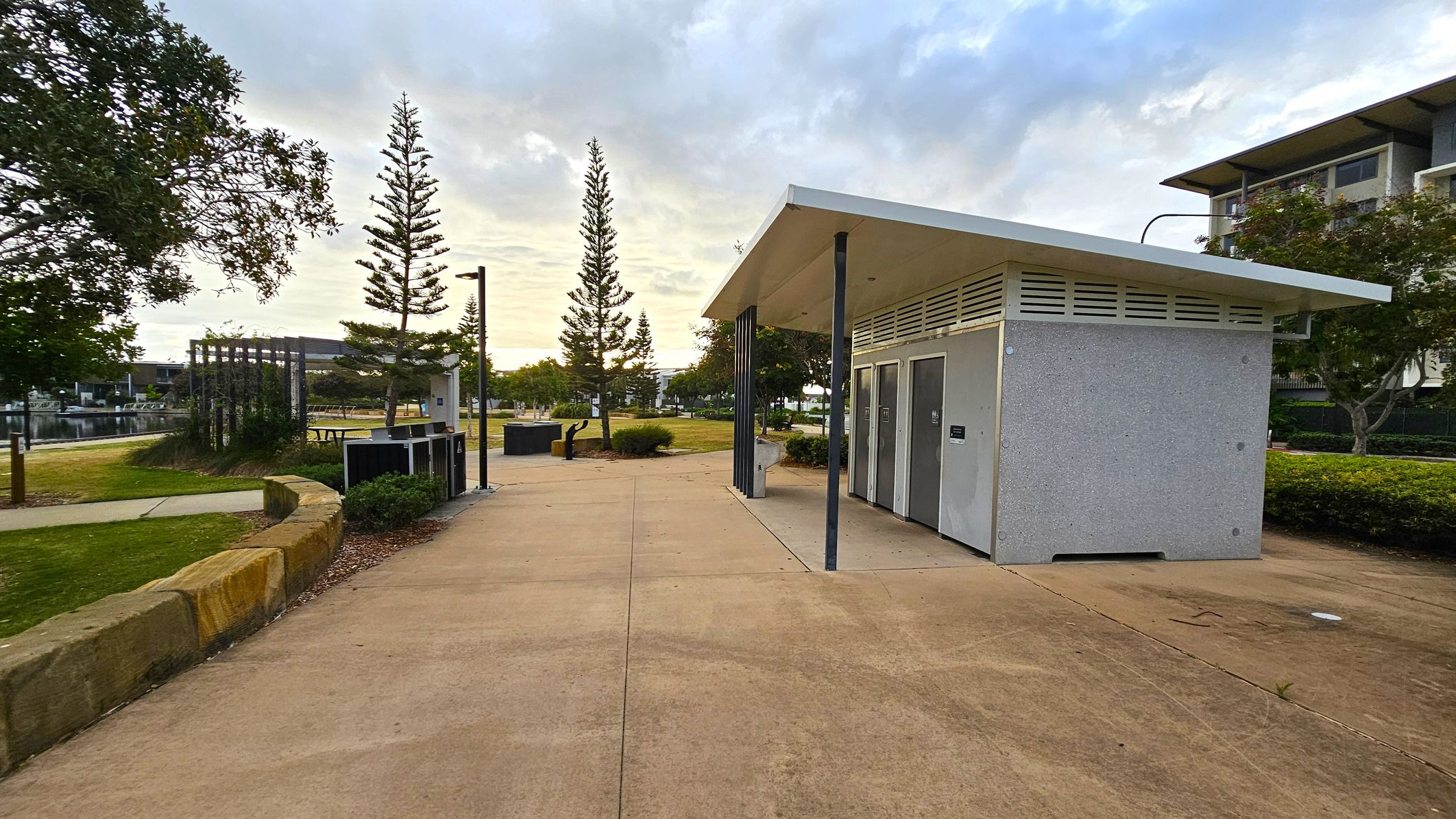 A public restroom building with a slanted roof is on the right, featuring grey doors and a textured exterior. The path in front is wide and paved. On the left, there are trees and a grassy area with a stone border. In the background, a body of water is visible, along with more trees and a modern building. The sky is overcast, and there are street lamps along the path.