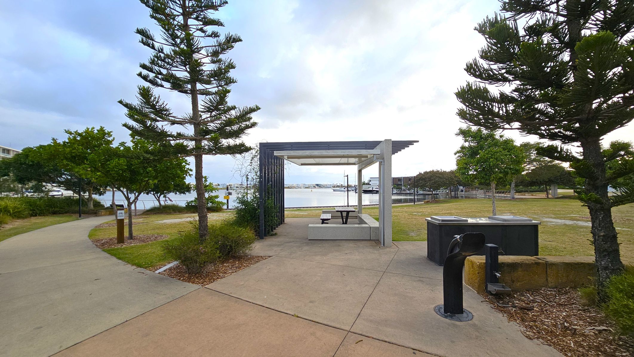 A park scene with a concrete path curving to the left, bordered by trees and shrubs. In the centre, a sheltered picnic area with a table and benches overlooks a body of water with boats. To the right, a black drinking fountain and a barbecue stand are visible. The ground is a mix of concrete and grass, with trees scattered throughout.