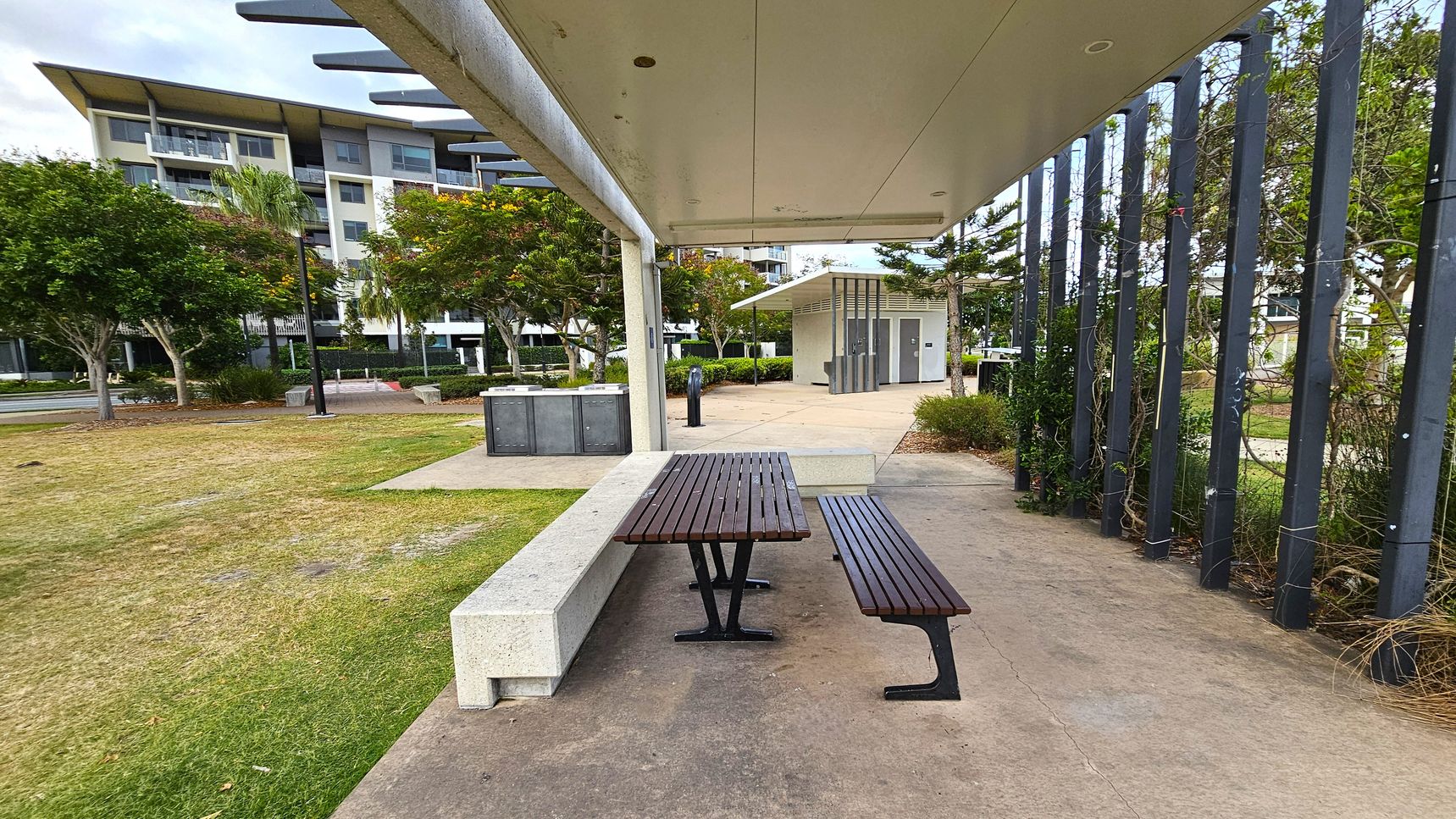A covered outdoor area with a wooden picnic table and bench on a concrete floor. To the right, vertical metal beams support climbing plants. In the background, a small building with doors is visible, surrounded by trees and bushes. On the left, a grassy area with more trees and a multi-storey building can be seen. The ceiling is flat and extends over the seating area.
