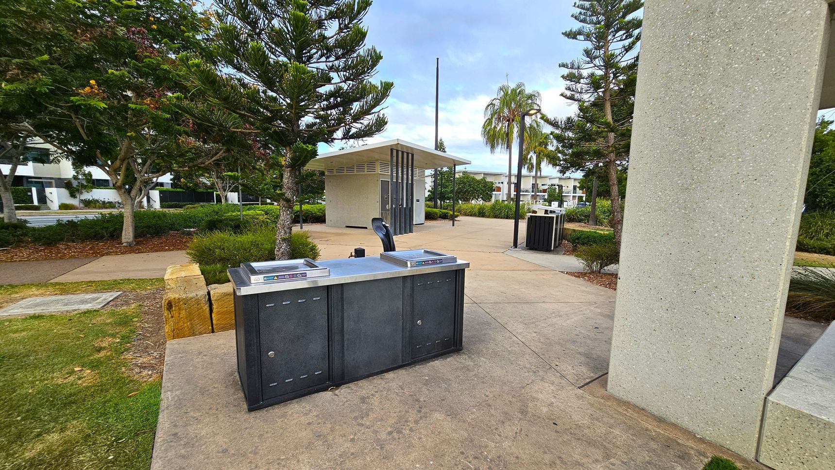 A public park area with a concrete path and a modern barbecue station in the foreground. To the left, there are trees and a grassy area with a stone bench. In the middle, a small building with a slanted roof is visible, likely a restroom or utility structure. To the right, a large concrete pillar is partially visible. In the background, more trees and a few buildings can be seen, along with a palm tree near the centre.