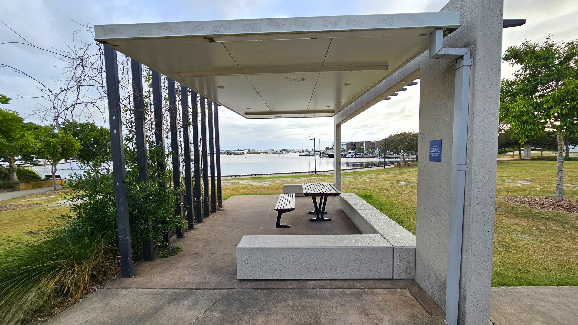 A sheltered picnic area with a metal roof and concrete supports. In the centre, there is a wooden picnic table with attached benches. The ground is a mix of concrete and grass. On the left, there are vertical metal bars with climbing plants. In the background, a body of water with boats and buildings is visible. On the right, there are trees and a grassy area.