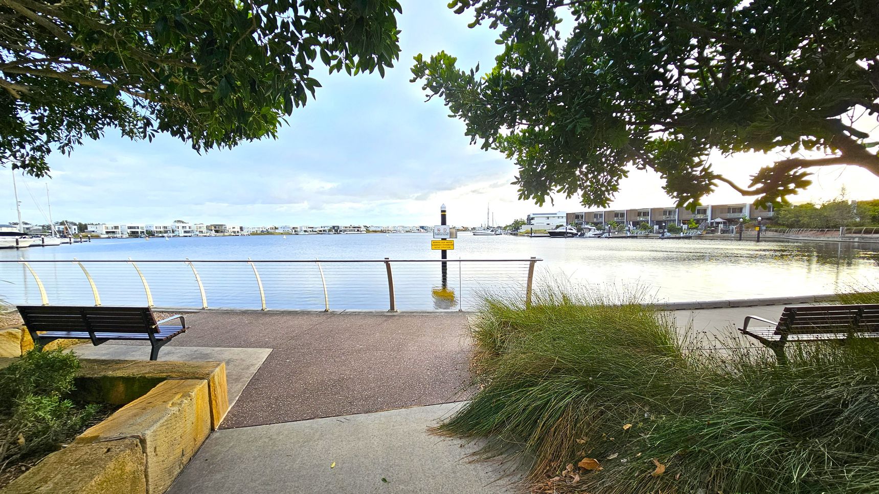 A waterfront scene with a paved path leading to a railing overlooking the water. Two benches are positioned on either side of the path, surrounded by grass and shrubs. Trees with dense foliage frame the top of the image. Across the water, there are modern buildings and boats docked at a marina. A sign is mounted on a post in the water near the centre. The area is calm and well-maintained.
