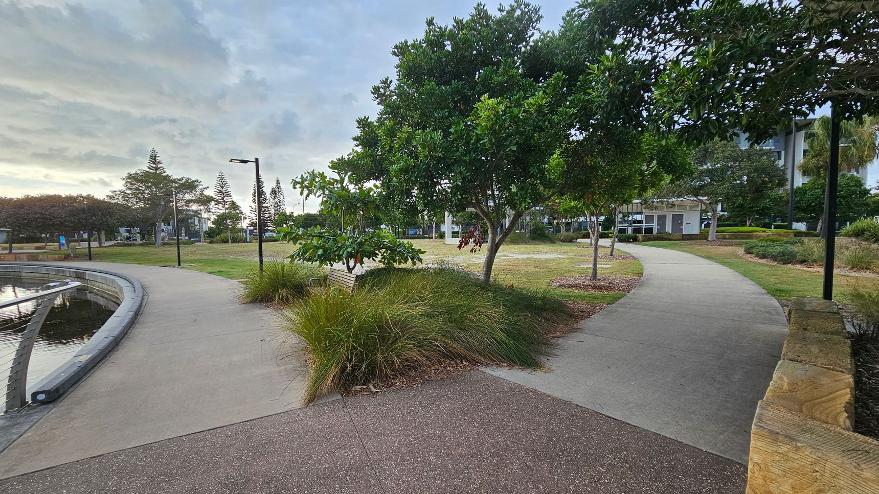 A park scene with two diverging concrete paths. The left path runs alongside a body of water with a low railing, while the right path curves through grassy areas and trees. In the centre, there are bushes and a bench beneath a tree. Several lamp posts line the paths. In the background, there are more trees and a building. The foreground features a reddish-brown textured surface and a wooden edge on the right.