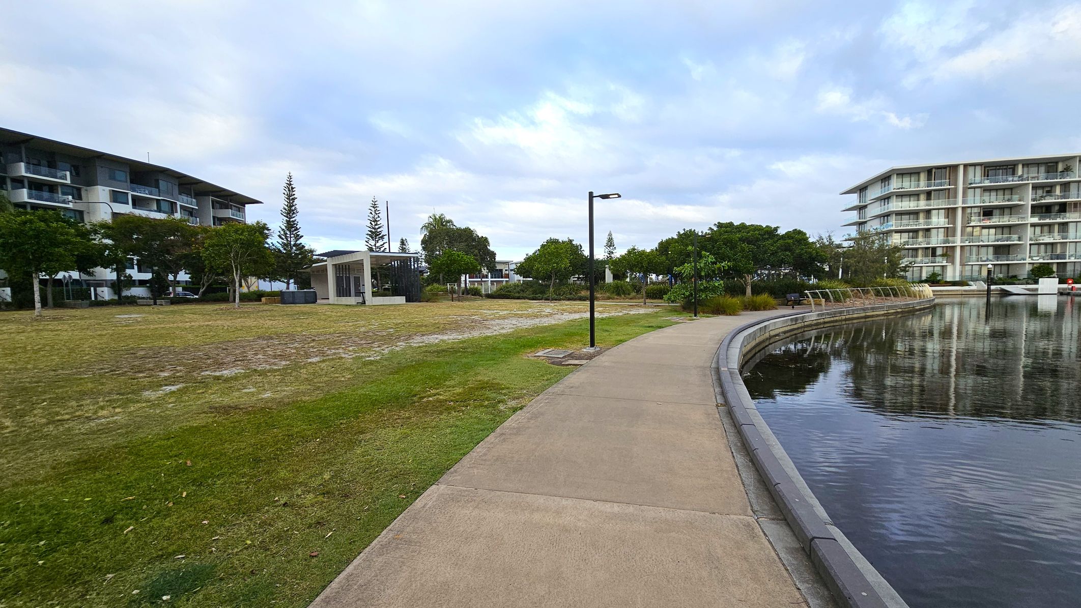 A curved pathway runs alongside a body of water on the right, with reflections of nearby buildings visible on the surface. On the left, there's a grassy area with scattered trees and a modern, open-sided shelter. Two multi-storey buildings are visible in the background, one on each side, with balconies and large windows. Street lamps line the path, and the area appears to be a landscaped urban park.