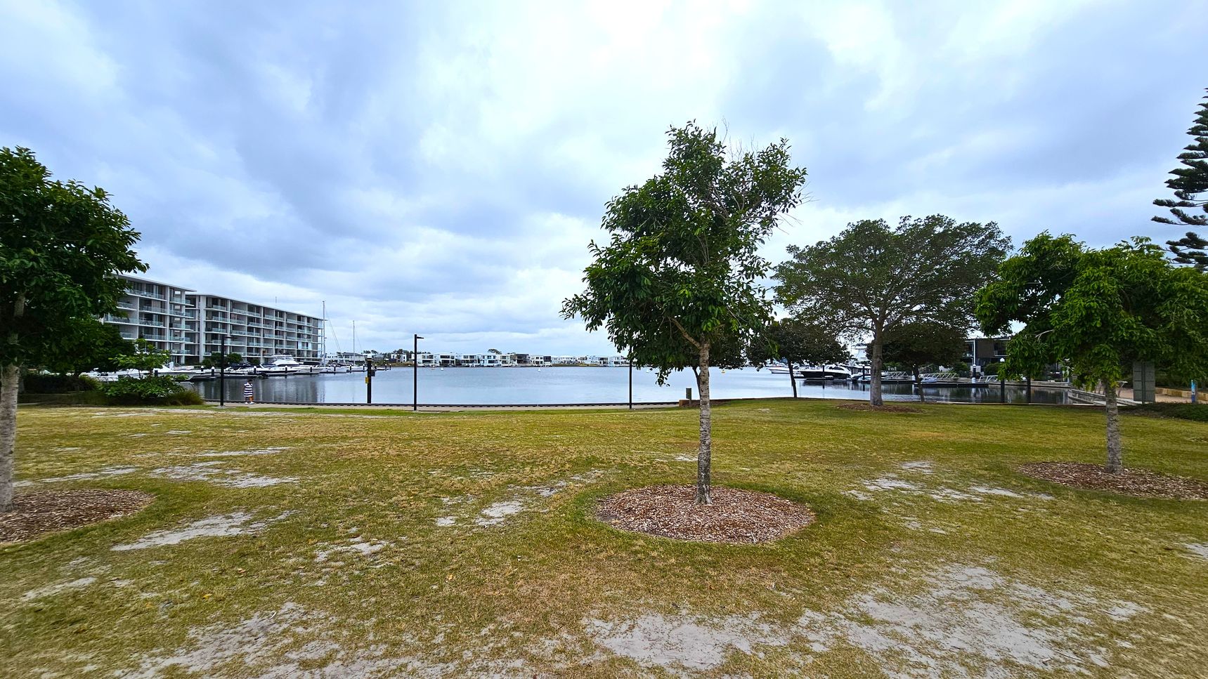 A grassy area with scattered trees in the foreground. In the background, a body of water is visible, bordered by a row of modern, multi-storey buildings on the left. Several boats are docked along the water's edge. On the right, more trees and a few structures are visible. The sky is overcast, casting a muted light over the scene.