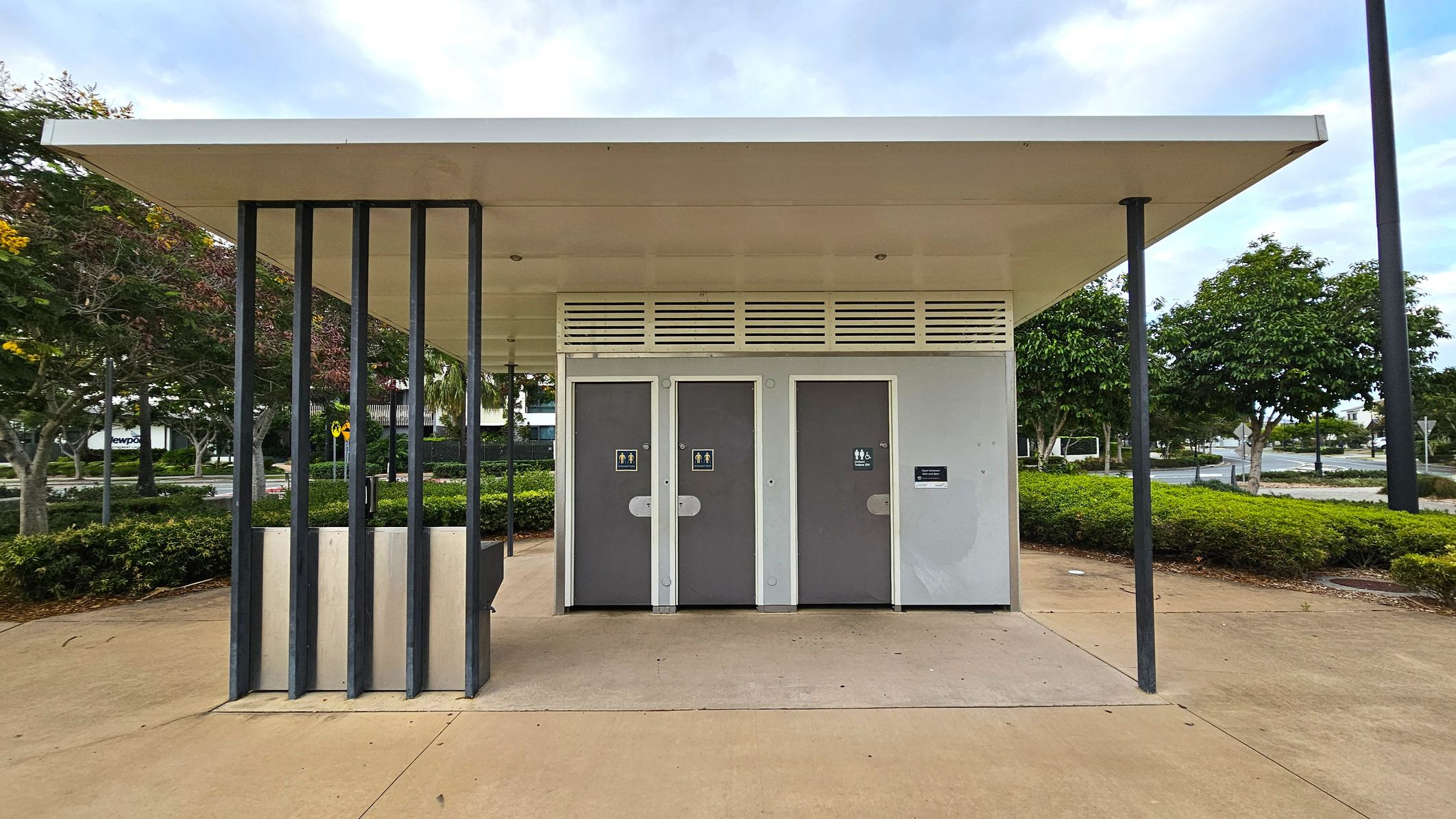 A public toilet block with a flat roof and three grey doors, each marked with gender-neutral signs. The structure has vertical black metal supports on the left. The ground is paved with light brown concrete. In the background, there are green bushes and trees with some red and yellow leaves. A road and a few parked cars are visible further back.