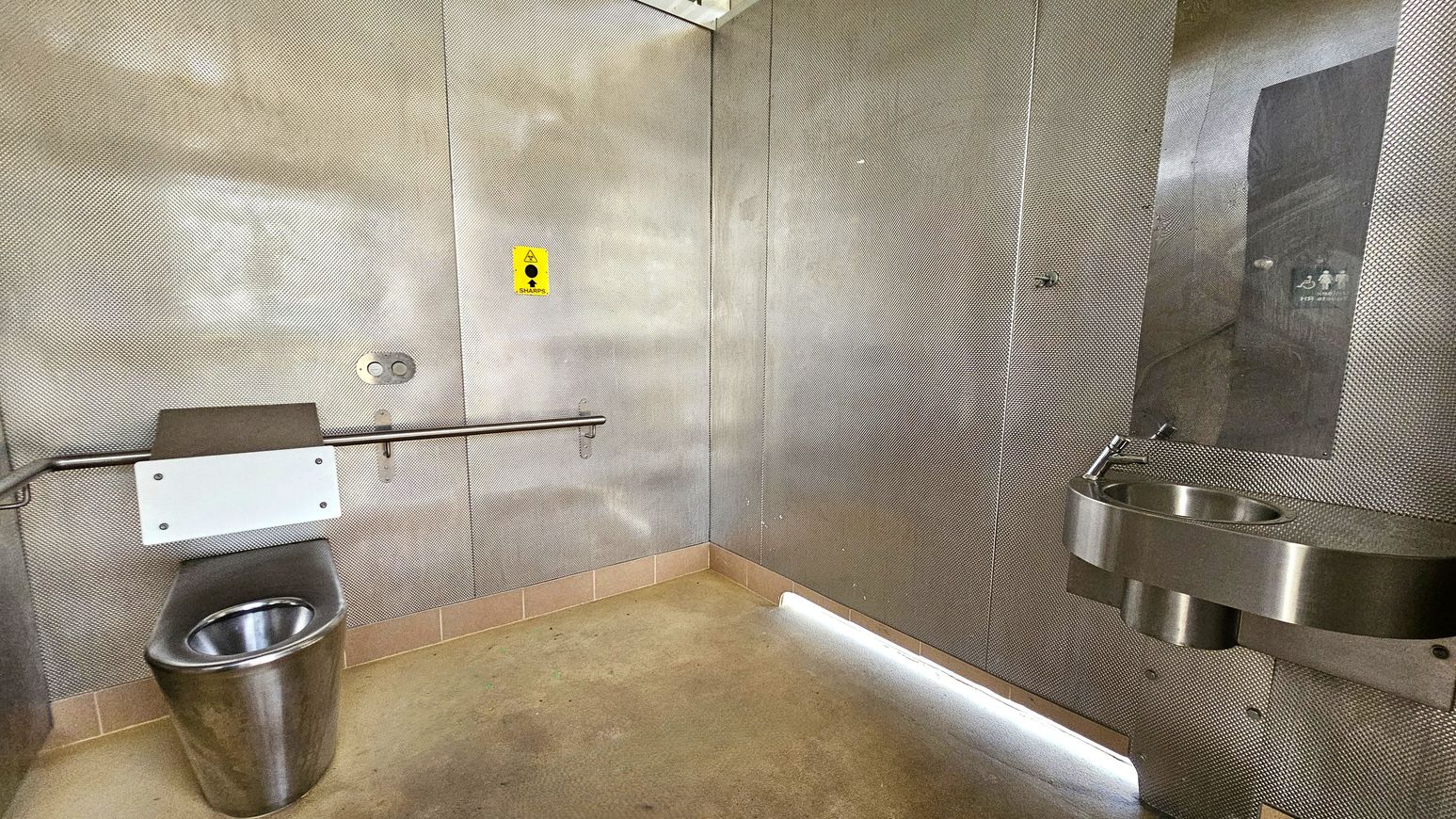 A public restroom with metallic walls and a concrete floor. On the left, there is a stainless steel toilet with a white panel and a horizontal grab bar. A yellow sharps disposal sign is visible above. On the right, a stainless steel sink is mounted on the wall with a small mirror above it. The ceiling is not visible.