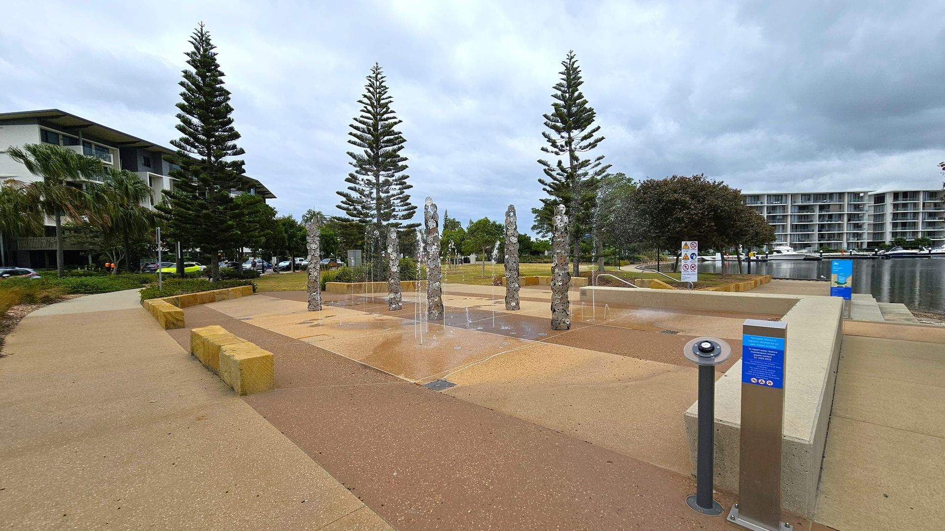 A public space with a water feature in the centre, consisting of several vertical stone columns with water jets. Surrounding the fountain are paved walkways and low stone benches. On the left, there are tall pine trees and a modern building with balconies. On the right, more trees and a multi-storey building are visible near a body of water with boats. The ground is a mix of light and dark paving, and there are informational signs near the fountain.