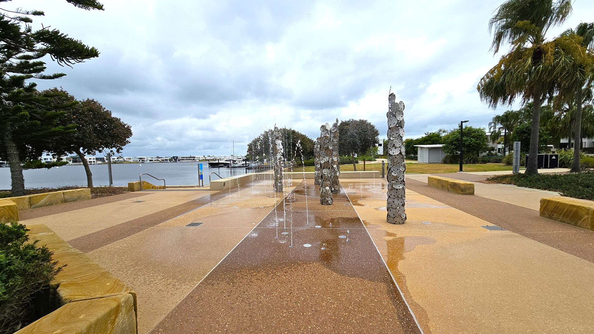 A waterfront area with a paved walkway featuring vertical sculptures made of circular metal pieces. Water jets are integrated into the ground, creating a fountain effect. On the left, there are trees and a view of the water with boats docked in the distance. On the right, more trees and a small building are visible. The ground is a mix of light and dark brown paving, and there are stone benches along the path.