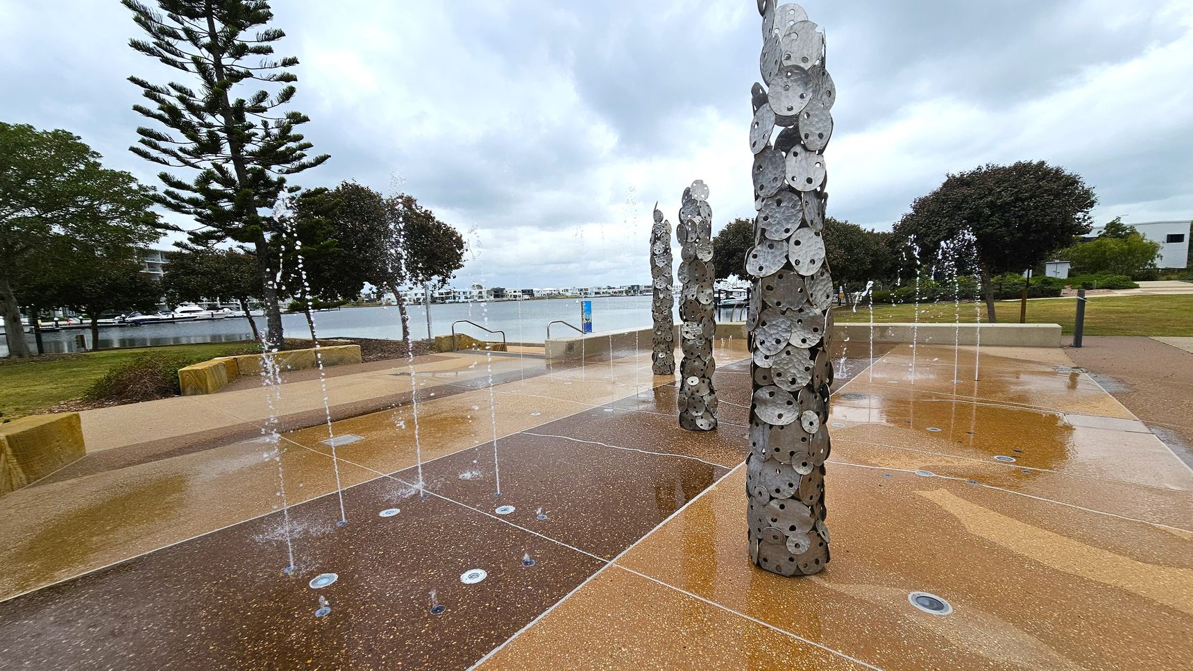 A waterfront area with a decorative fountain in the foreground, featuring vertical columns adorned with circular metal discs. Water jets are spraying upwards from the ground. To the left, a tall pine tree and other greenery are visible. In the background, a body of water with moored boats and a line of modern buildings can be seen. The ground is a mix of textured concrete and stone, with some wet patches. The sky is overcast.