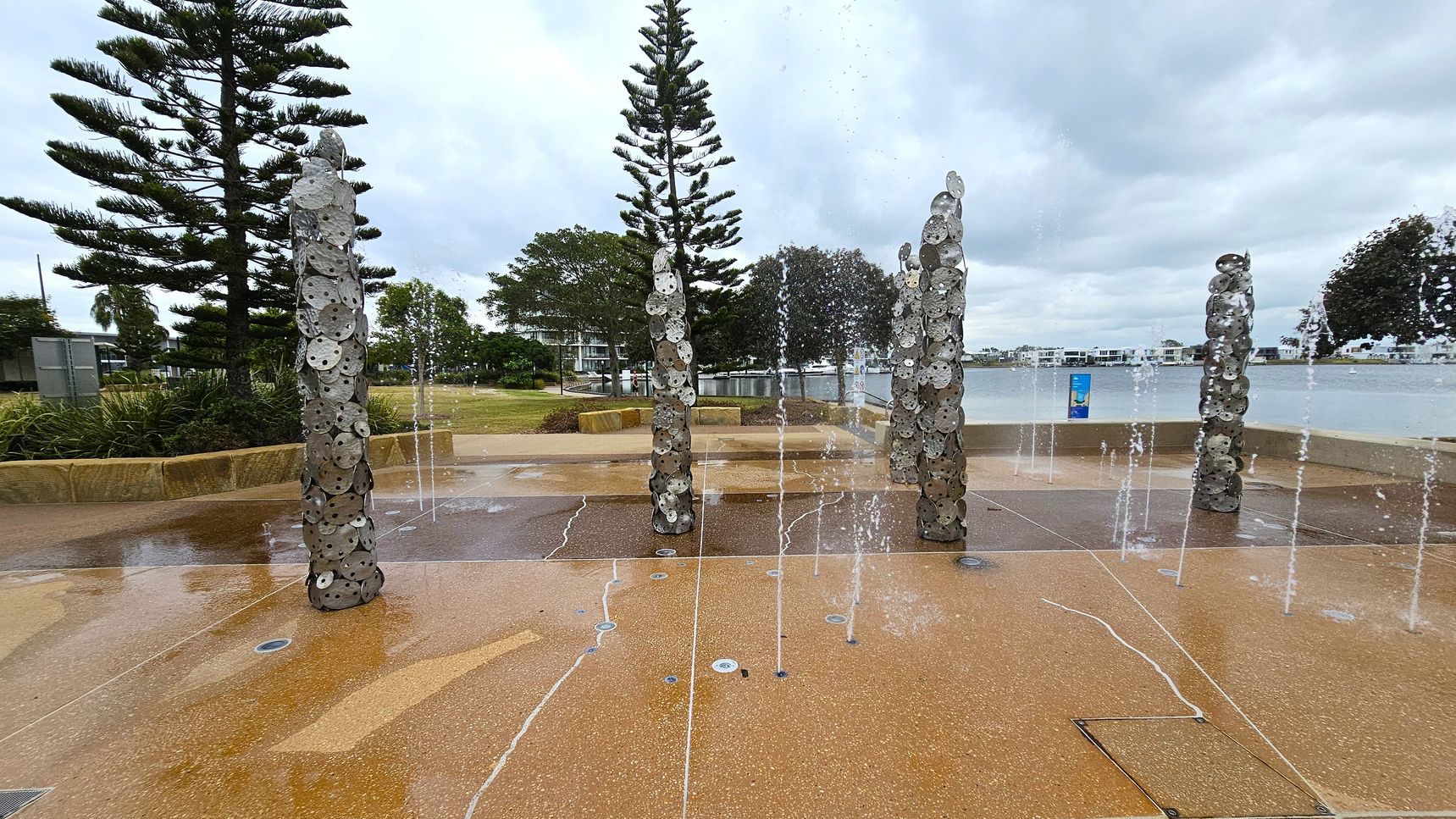A paved area with water jets shooting upwards from the ground. In the centre, there are several vertical sculptures made of circular metal discs stacked on top of each other. Surrounding the area are tall pine trees and other greenery. In the background, there's a body of water with buildings visible on the opposite shore. The sky is overcast, and the ground appears wet, reflecting the water from the jets.