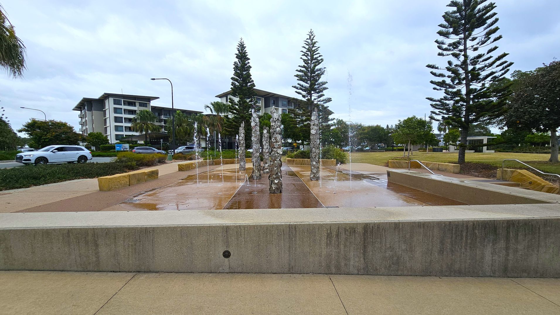 A water feature with vertical jets and decorative stone columns is in the foreground, surrounded by a paved area. In the background, there are several tall pine trees and a modern multi-storey building. To the left, a white car and a grey car are parked on a road lined with greenery. The area is landscaped with trees and shrubs, and there are stone benches and pathways. The sky is overcast.