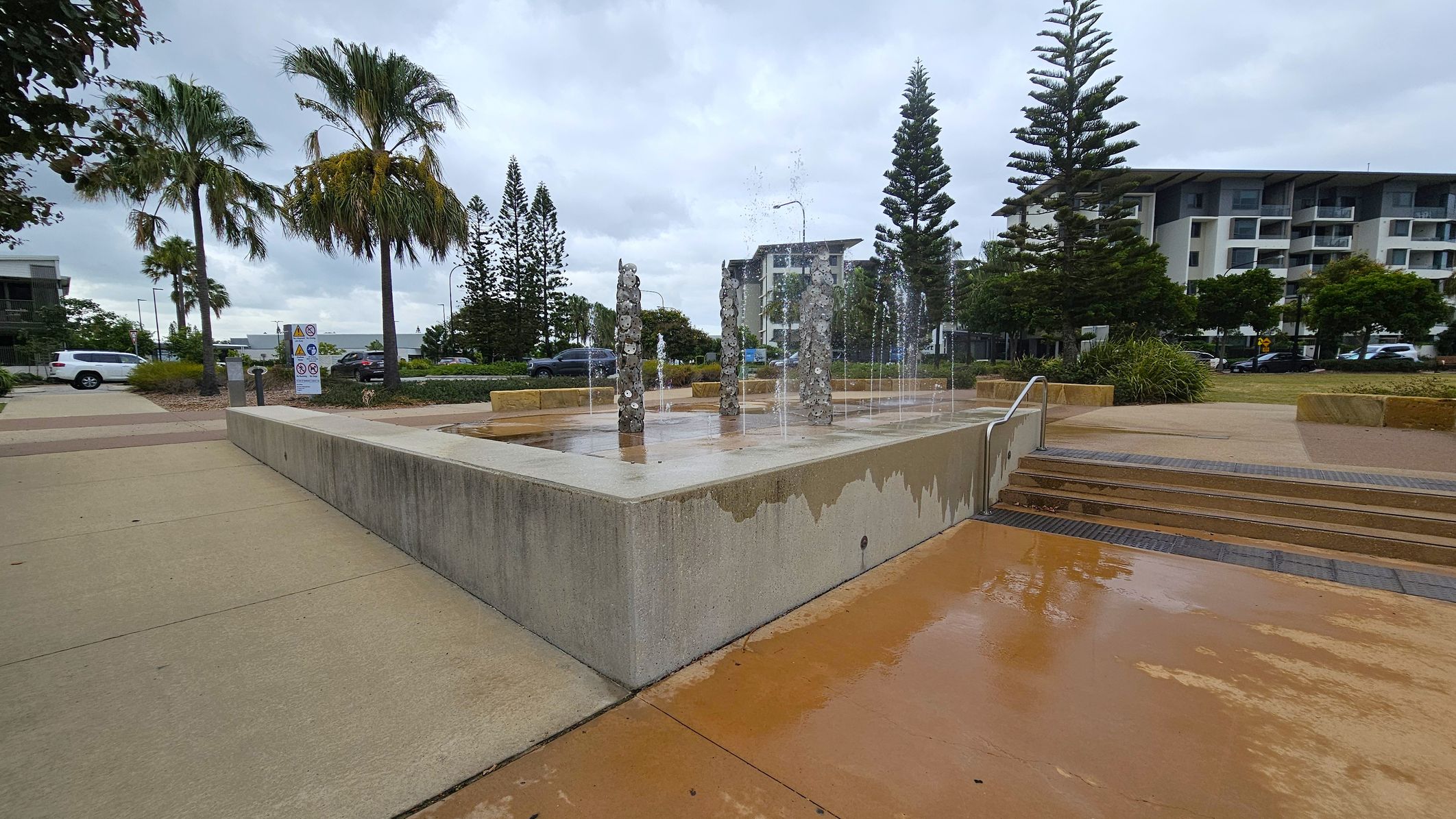 A concrete fountain with water jets is in the centre, surrounded by a low wall. On the left, there are palm trees and a sign with warnings. A white vehicle is parked nearby. On the right, steps lead up to the fountain area, with a large building and more trees in the background. The ground is wet, and the sky is overcast.
