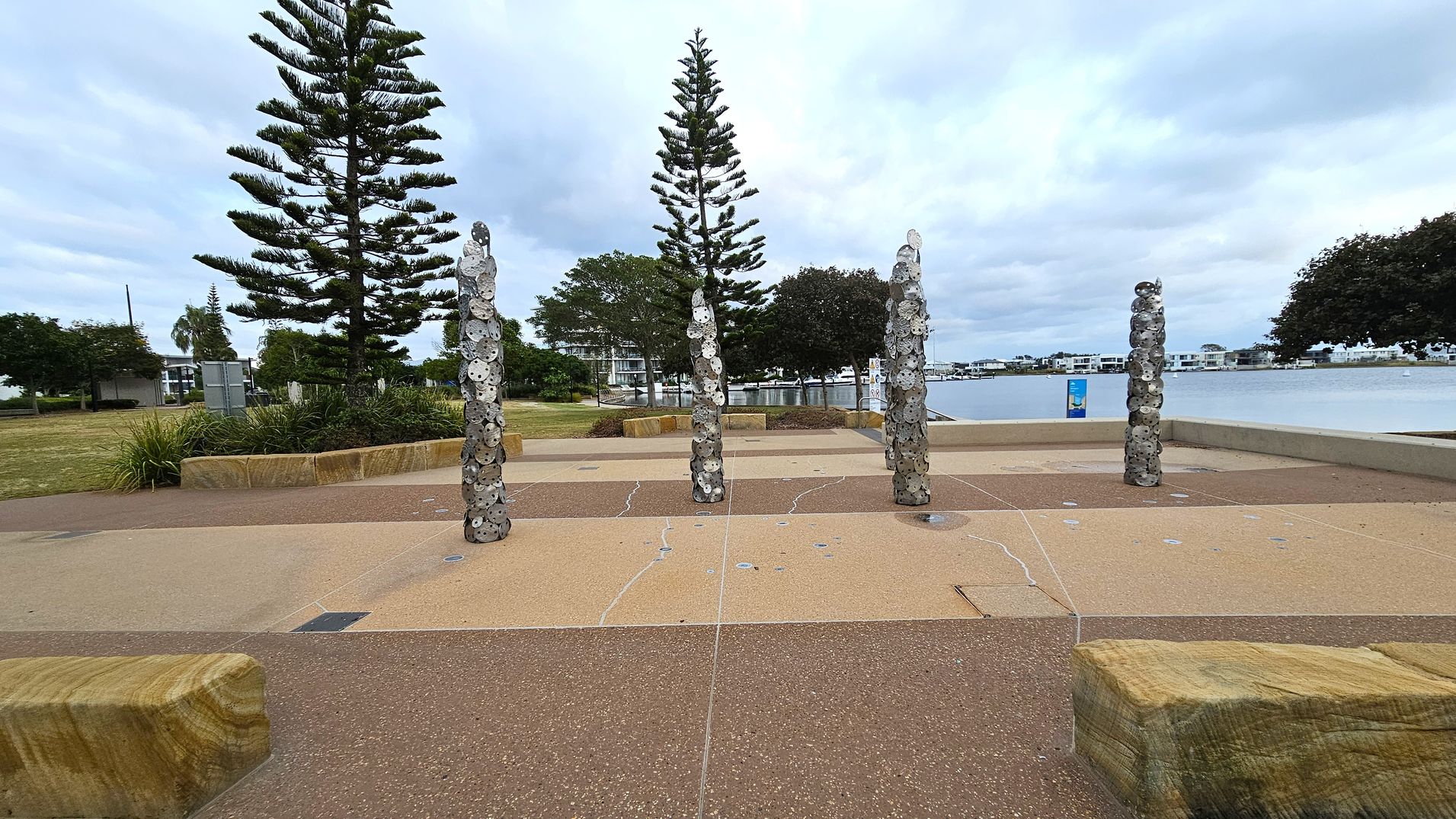 Five vertical sculptures made of stacked circular metal pieces are arranged in a row on a paved area. Behind them, two tall, symmetrical pine trees stand prominently. To the left, there is a grassy area with more trees and a building in the distance. On the right, a body of water is visible, bordered by modern buildings. The ground is a mix of textured and smooth surfaces, with stone benches placed around the area.