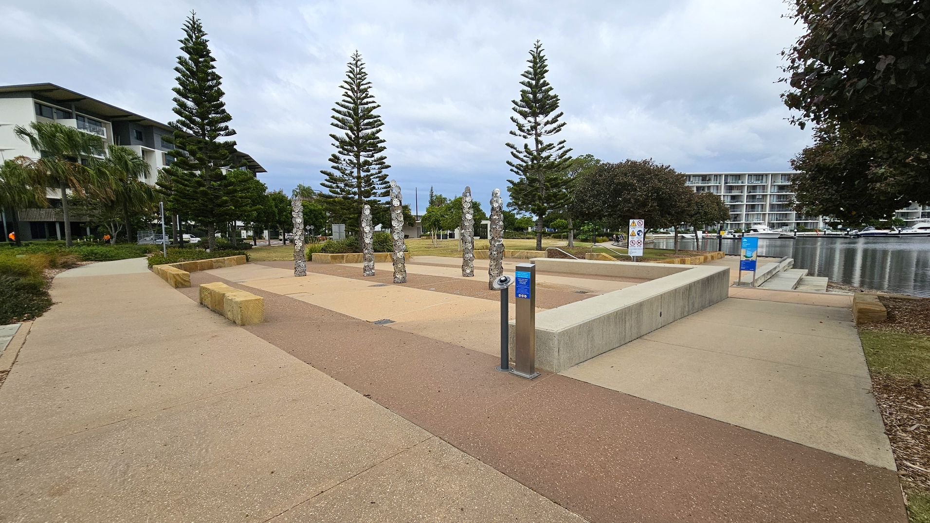 A paved area with a series of vertical stone sculptures in the centre. On the left, there are tall pine trees and a modern building with large windows. On the right, more trees and a body of water with boats docked alongside a multi-storey building. The ground is a mix of beige and reddish-brown paving, with a metal post featuring a blue sign. The area is bordered by low stone walls and benches.