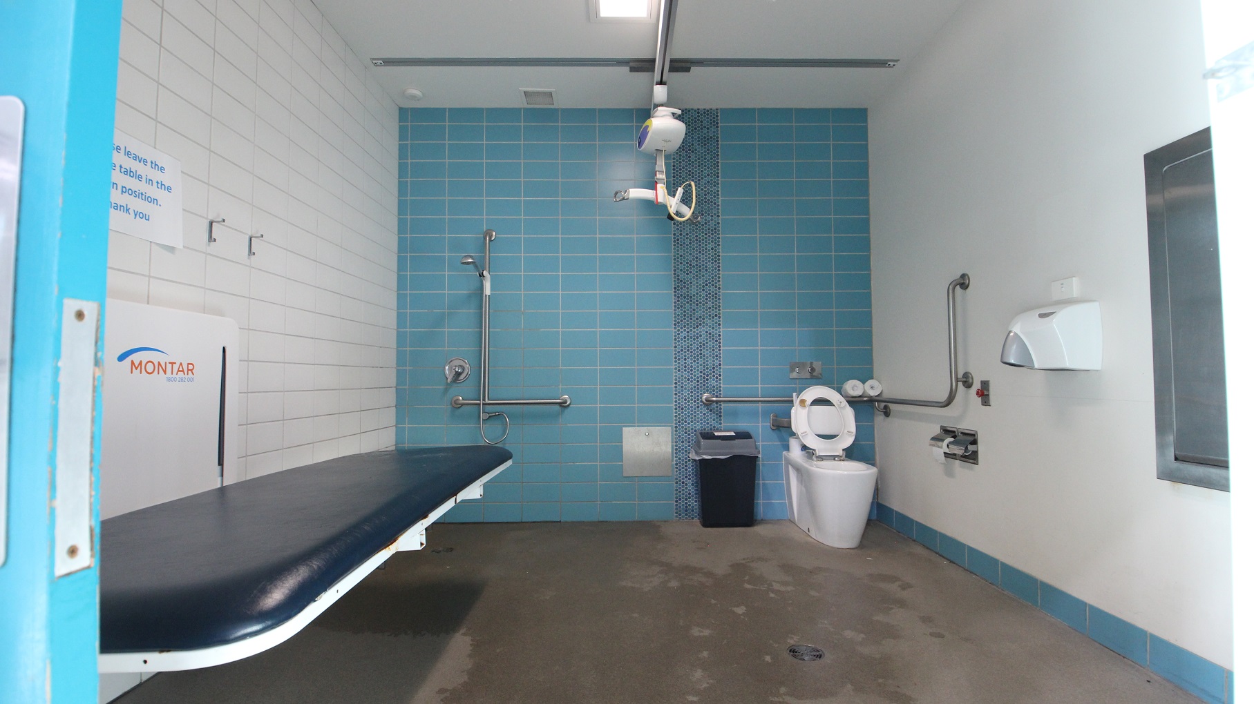 Inside the accessible bathrooms at Splash Devonport is a hoist, a dark blue cushioned adult change table, a toilet, a shower, and guard rails. Beside the toilet bowl is the rubbish bin. The back wall is made of blue tiles, the left is white tiles, while the right is a smooth white wall. Inside the accessible bathrooms at Splash Devonport is a hoist, a dark blue cushioned adult change table, a toilet, a shower, and guard rails. Beside the toilet bowl is the rubbish bin. The back wall is made of blue tiles, the left is white tiles, while the right is a smooth white wall.