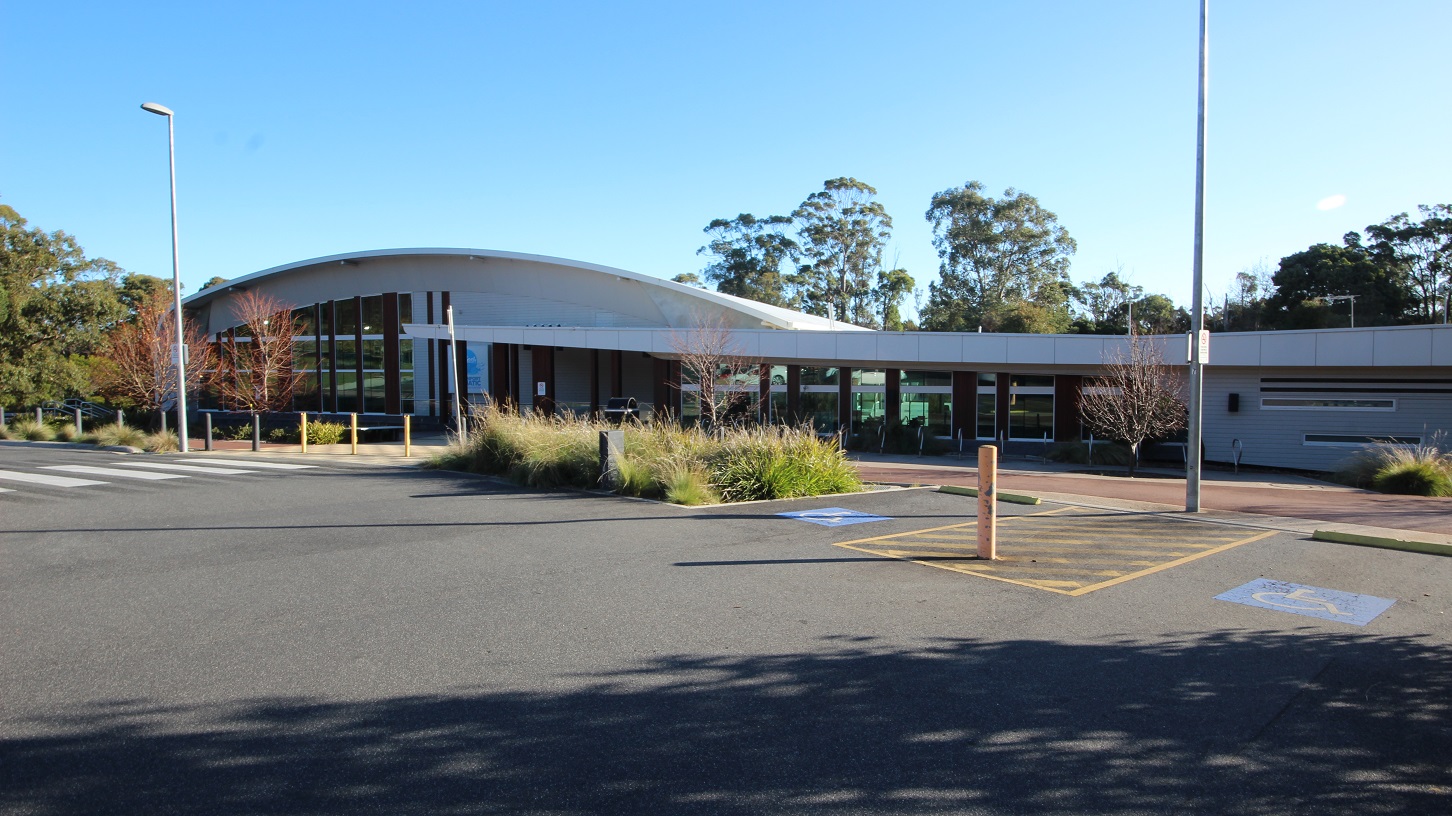 There is a pedestrian lane leading toward a fully accessible entrance-style ramp in Splash Devonport, and on the right is a plant box containing several bushes with moderate leaf length. Street lamp posts are scattered in the accessible parking spaces, distinguished by the persons with blue and white accessible parking signs, yellow bollards, and yellow lines. There is a pedestrian lane leading toward a fully accessible entrance-style ramp in Splash Devonport, and on the right is a plant box containing several bushes with moderate leaf length. Street lamp posts are scattered in the accessible parking spaces, distinguished by the persons with blue and white accessible parking signs, yellow bollards, and yellow lines.