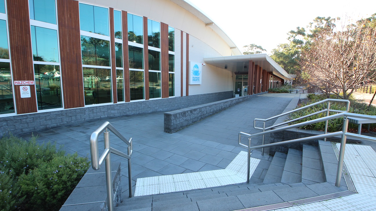 Staircase with metal handrails leading to one of the entrances of Splash Devonport. The floor is made of grey, square tiles. The walls of Splash Devonport are a combination of grey and white bricks, glass, and long stretches of wood arranged vertically. The glass walls reflect the car spaces and the surrounding area of the venue. Staircase with metal handrails leading to one of the entrances of Splash Devonport. The floor is made of grey, square tiles. The walls of Splash Devonport are a combination of grey and white bricks, glass, and long stretches of wood arranged vertically. The glass walls reflect the car spaces and the surrounding area of the venue.